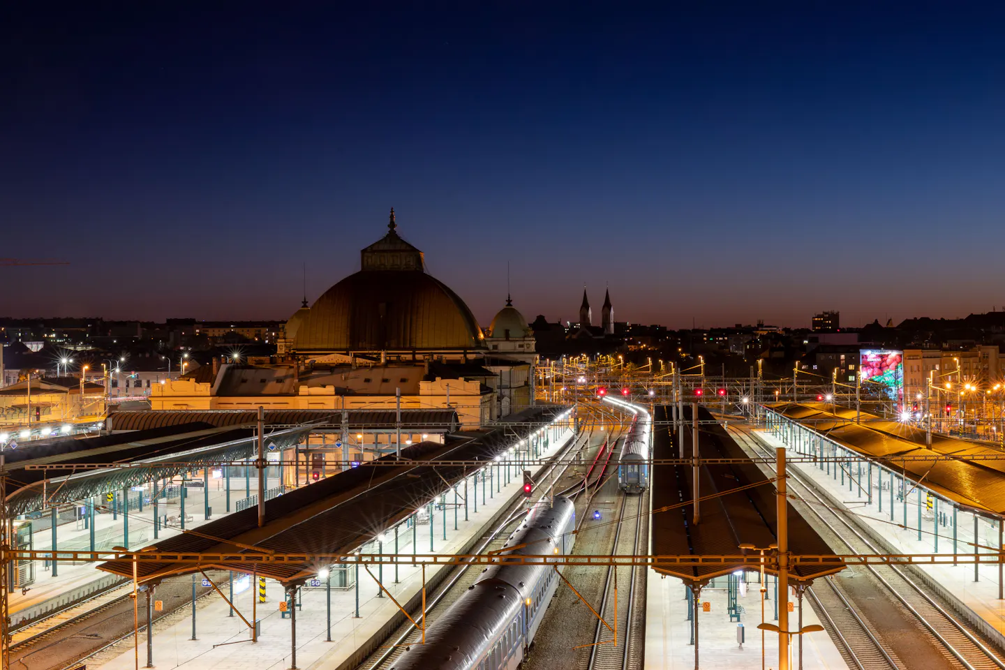 A brightly lit railway station at dusk with trains at several platforms below a domed building