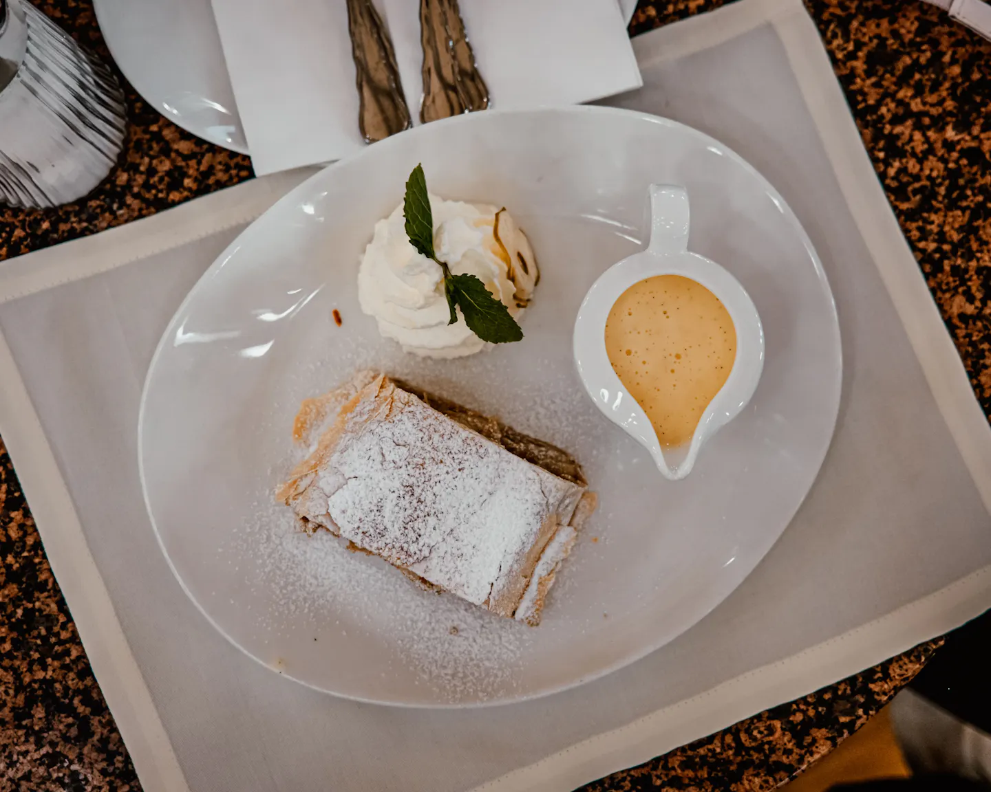 A sugar-dusted slice of apple strudel with whipped cream and a jug of vanilla sauce