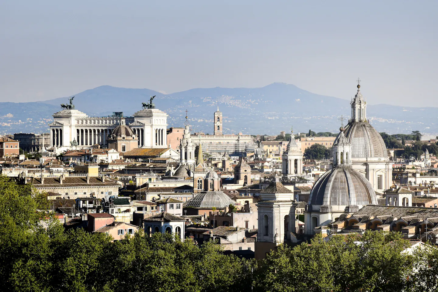 Rome's skyline of church domes and bell towers with the white Vittoriano on the left and the Alban Hills beyond, above a band of green trees