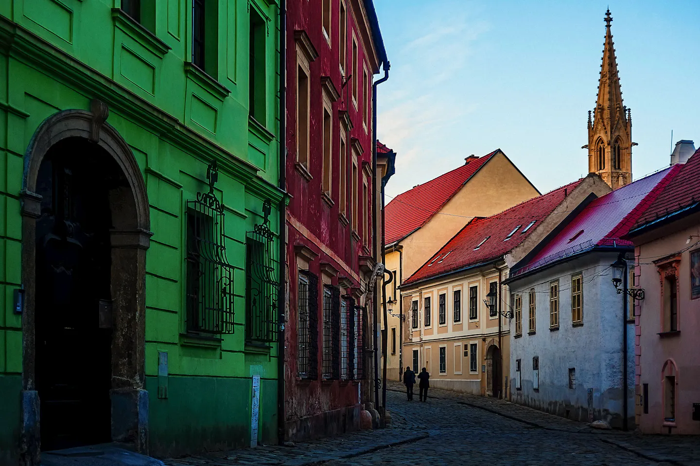 A cobbled Bratislava old-town street of green and red houses leading towards a Gothic spire