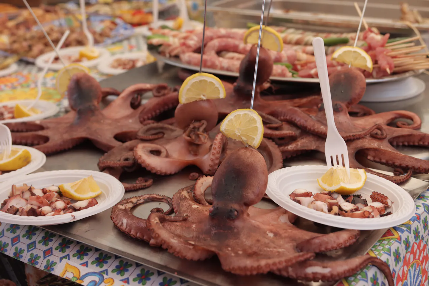 Boiled octopus with lemon wedges on a Sicilian street-food stall beside trays of skewers