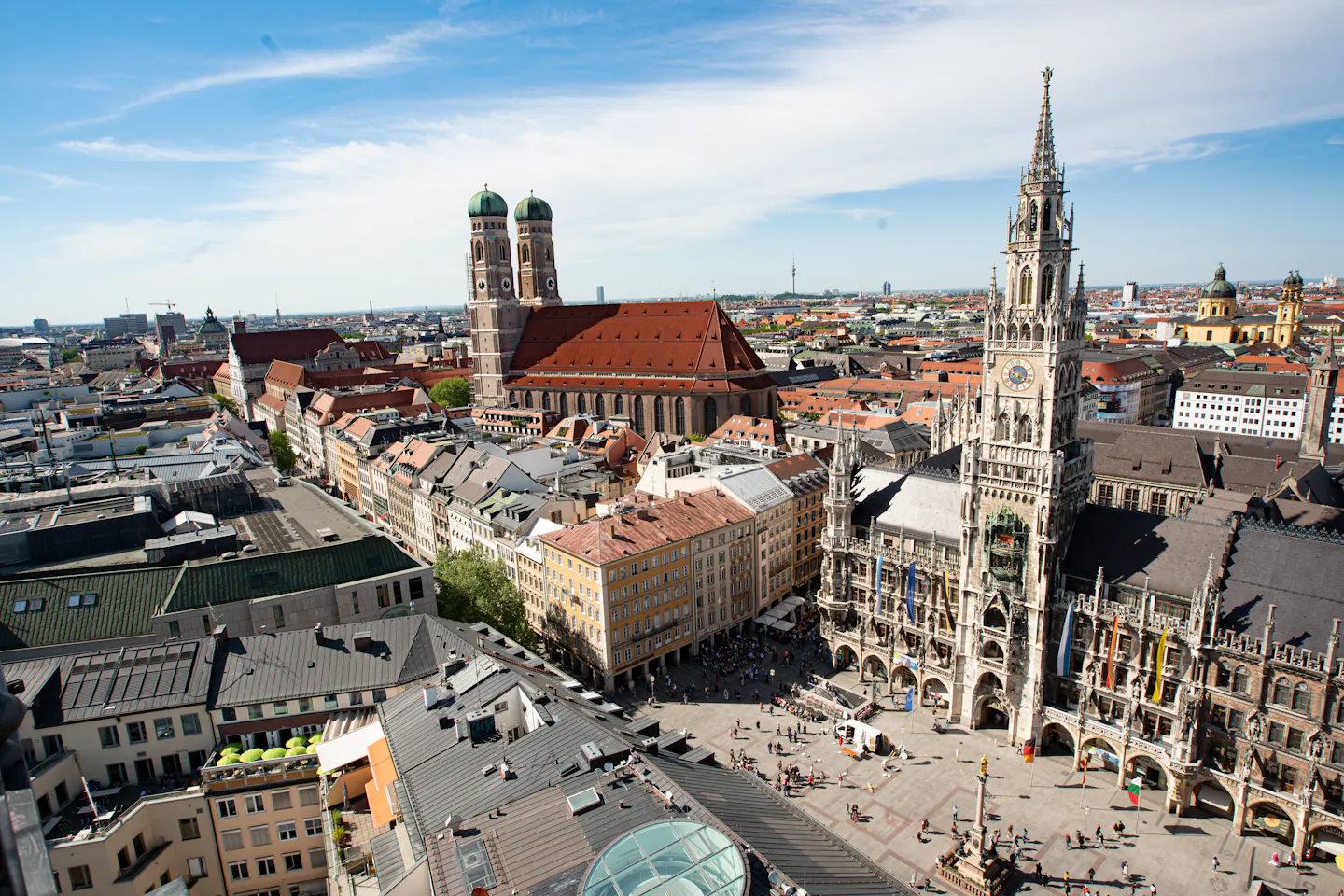 Munich’s Marienplatz from above, the spire of the New Town Hall beside the green onion domes of the Frauenkirche under a bright sky