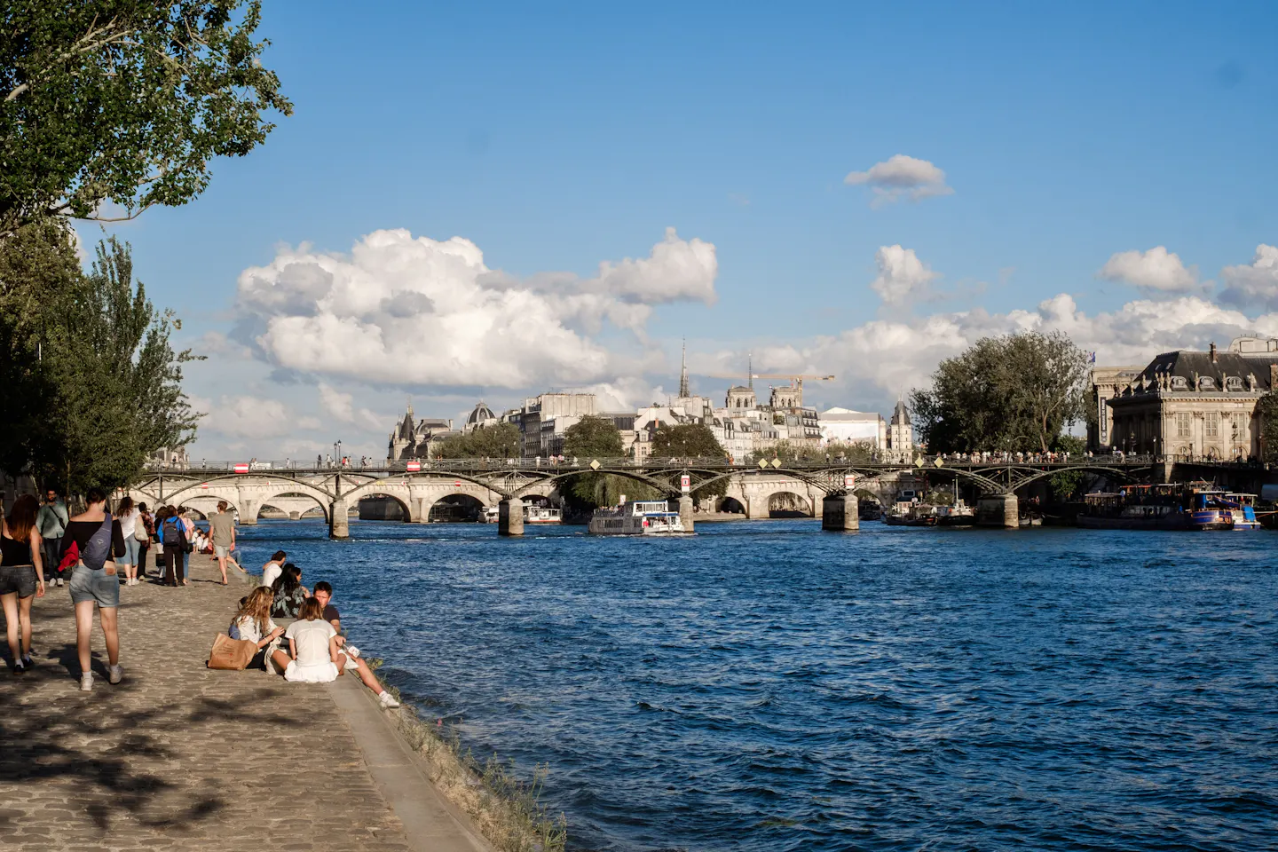 People sitting along the Seine riverbank on a sunny summer day near the Pont des Arts