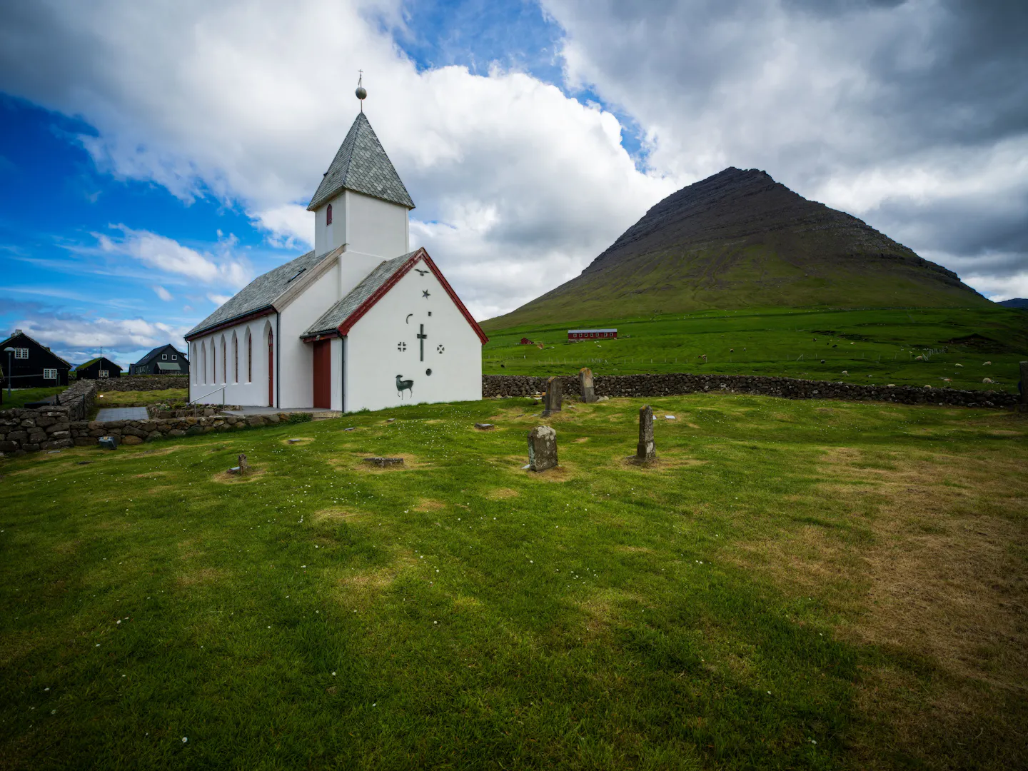 A small white Faroese church and its stone-walled graveyard on bright green grass below a steep pyramid-shaped mountain
