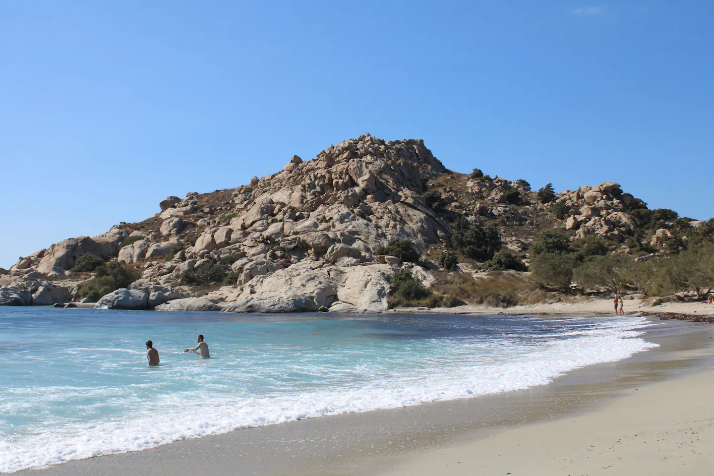 Turquoise water and white surf breaking onto a wide sandy beach below a boulder-strewn granite headland on a Greek island