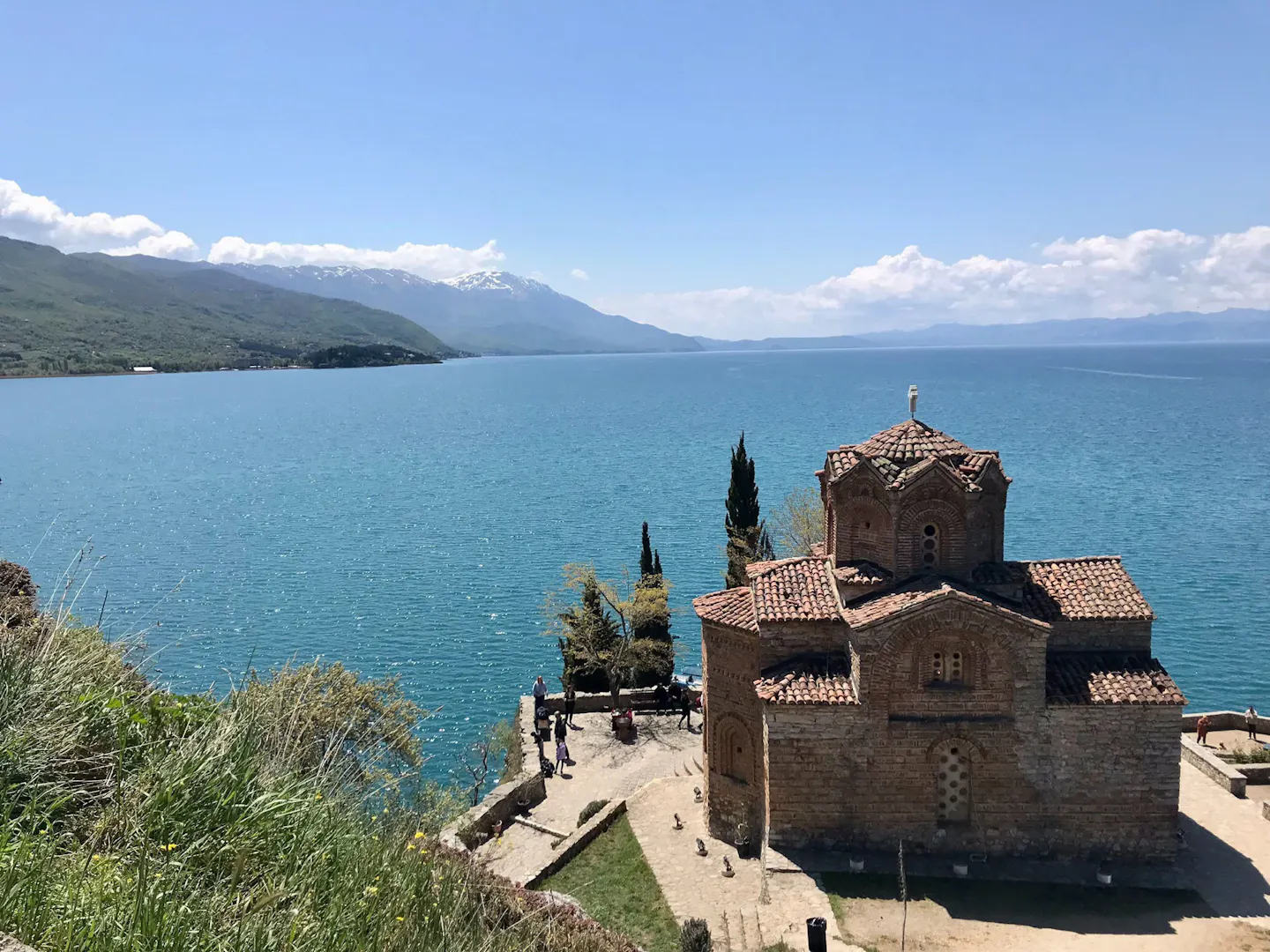 A small Byzantine stone-and-brick church on a headland above the turquoise water of Lake Ohrid, with snow-capped mountains along the far shore