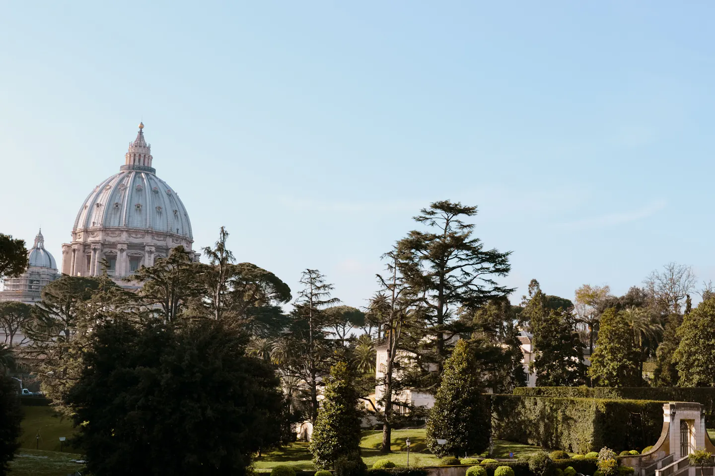 The dome of St Peter's Basilica rising pale above dark umbrella pines, cypresses and clipped hedges of a formal Roman garden under a clear winter-blue sky