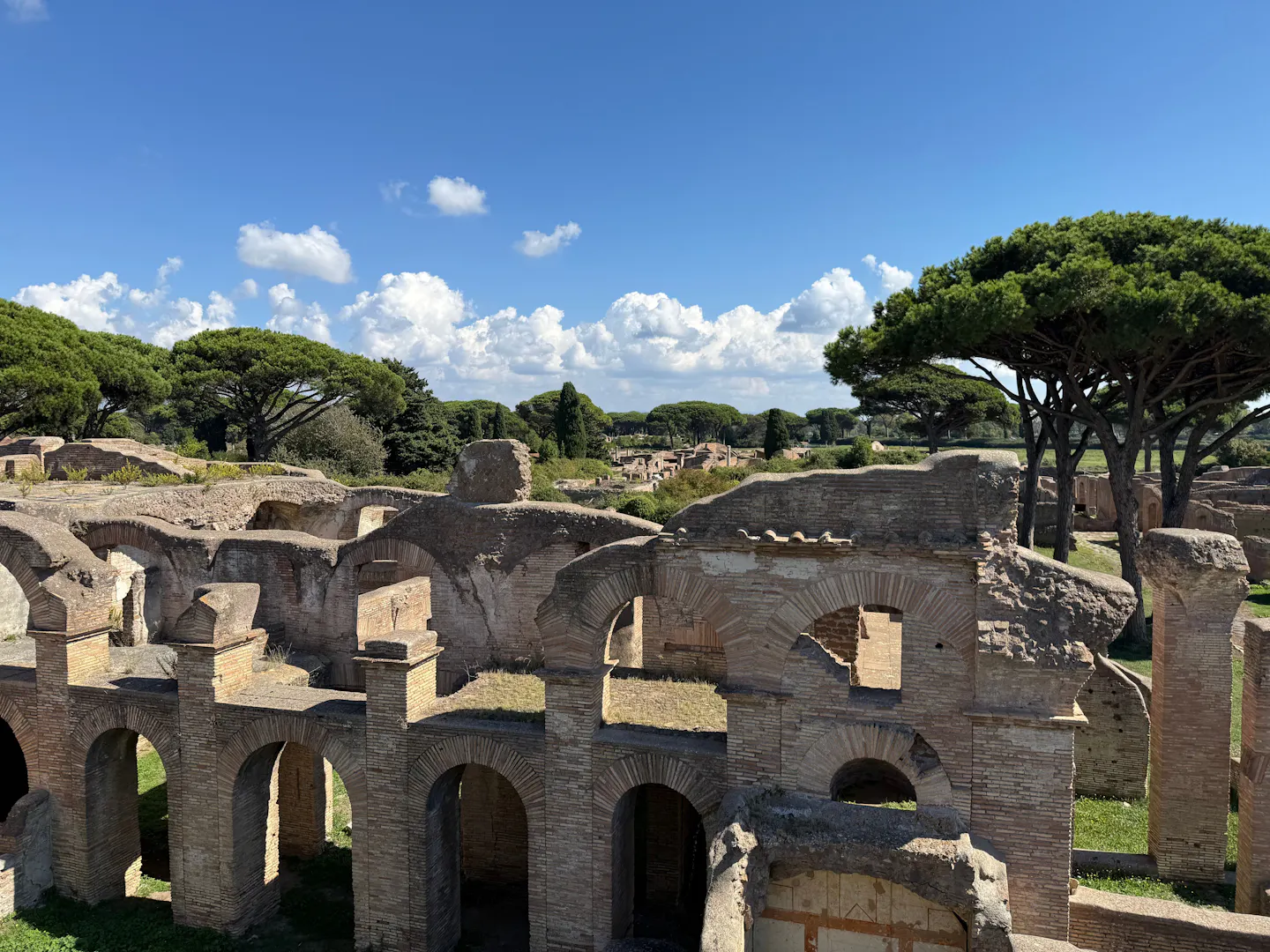 Rows of brick arches and roofless rooms in the Roman ruins of Ostia Antica, with umbrella pines and cypresses along the horizon under a deep blue sky and white cumulus