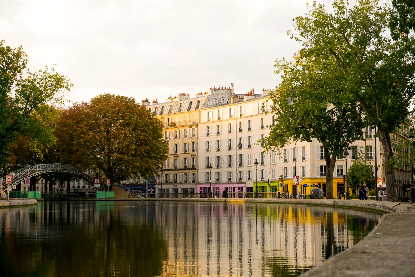 An iron footbridge over Canal Saint-Martin with facades and trees reflected in the water at golden hour