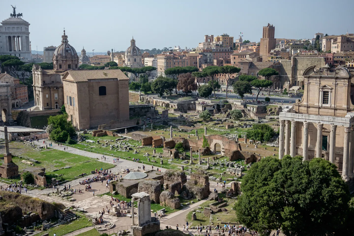 The ruins of the Roman Forum seen from above in bright sun, with the Temple of Antoninus and Faustina on the right, church domes and the Vittoriano on the skyline behind