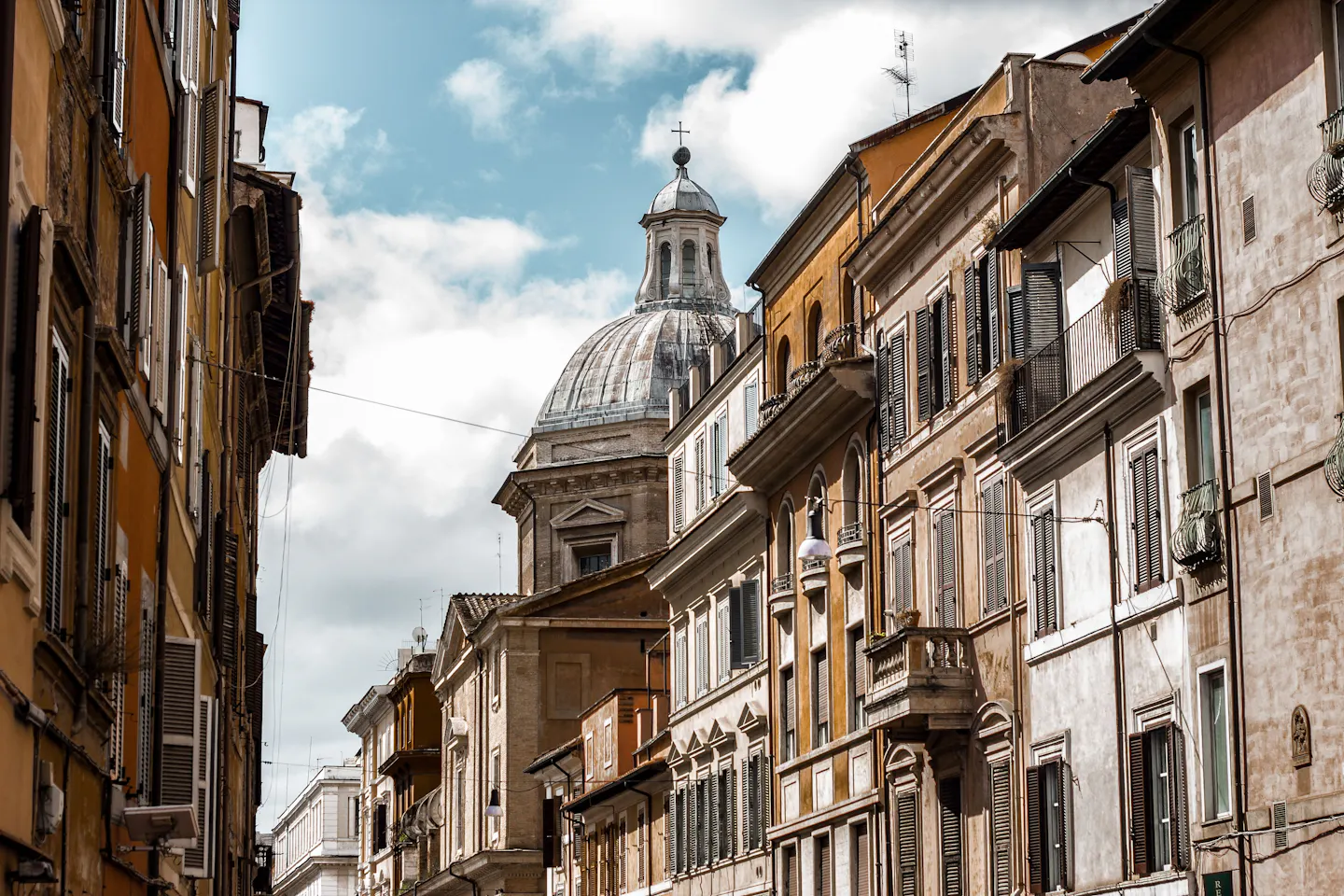 A sunlit central Rome street lined with tall ochre and cream palazzi, their shuttered windows fronted by rows of small wrought-iron balconies, with a church dome behind and big white clouds overhead