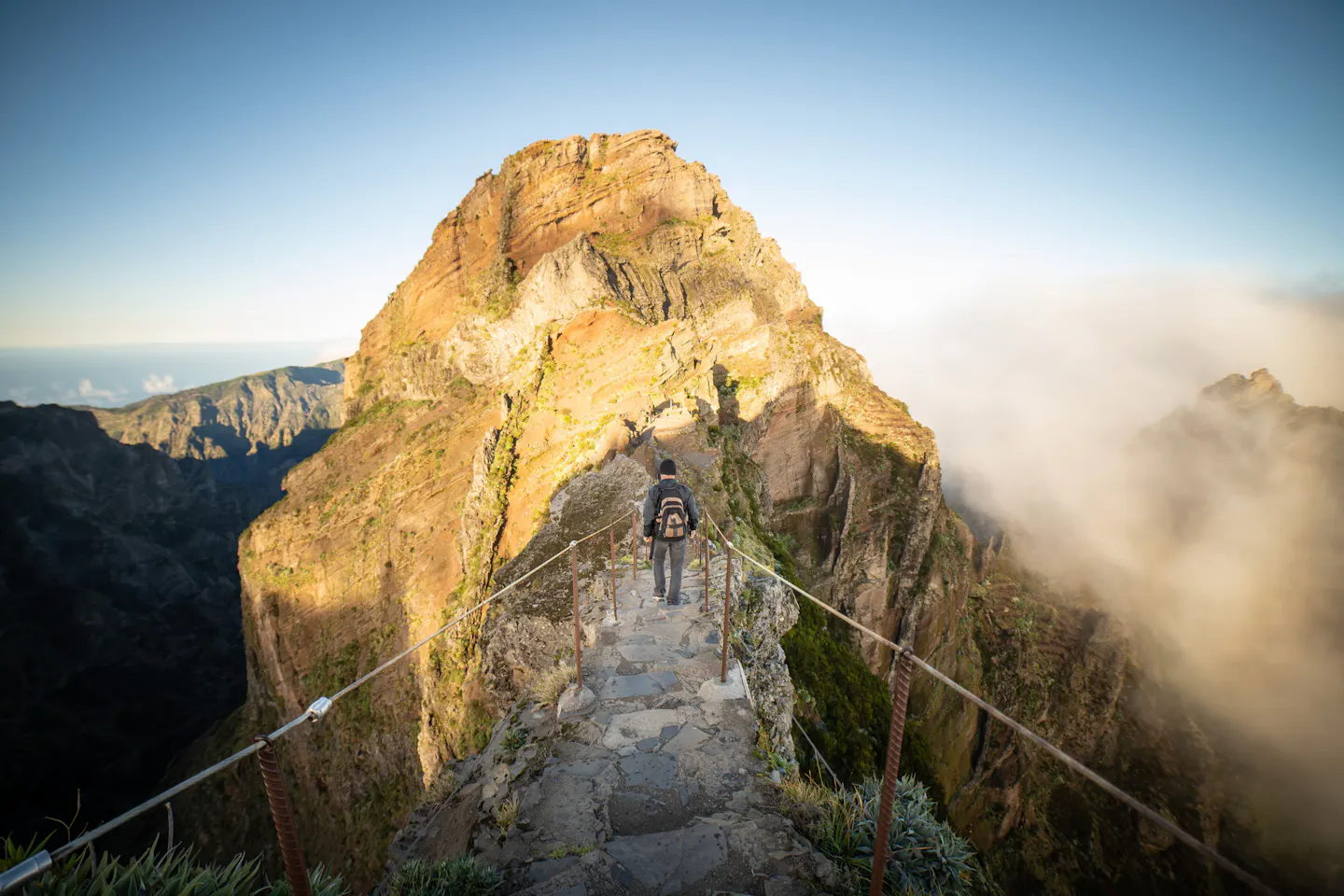 A stone stairway trail running along a narrow mountain ridge in Madeira, with a lone hiker walking towards a sunlit peak and a sea of cloud rolling in
