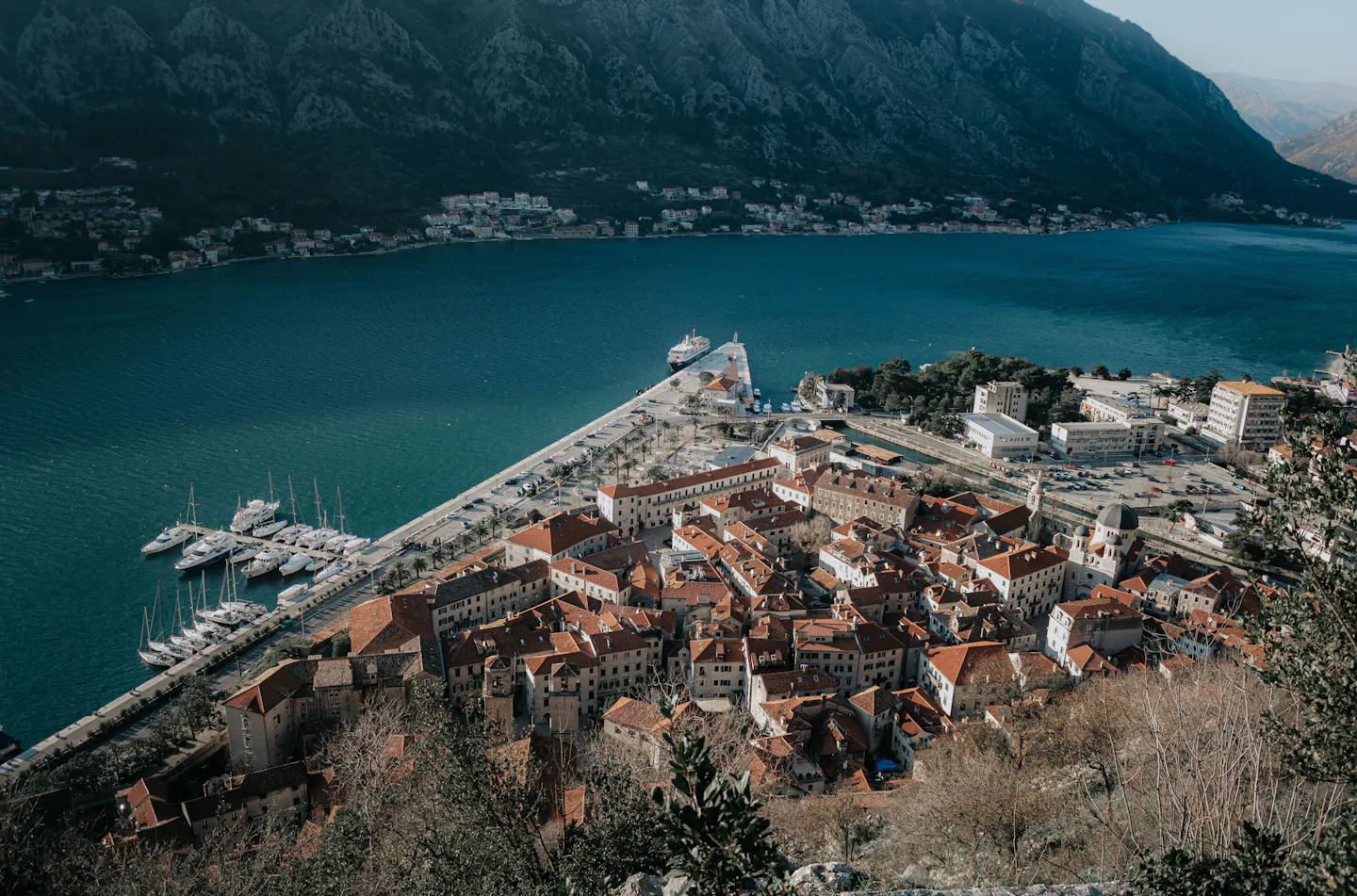 Kotor's walled old town of red-tiled roofs wedged between the deep teal Bay of Kotor and a sheer grey mountain wall, seen from the fortress trail above