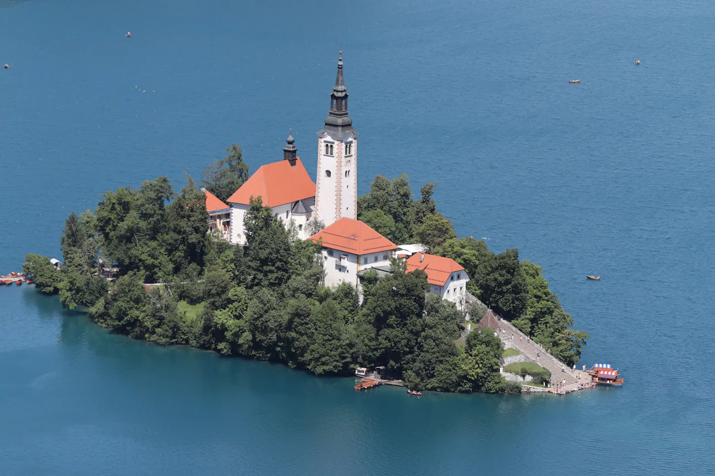 Bled Island seen from above, its white bell tower and orange-roofed church rising from dense green trees in the deep blue water of Lake Bled