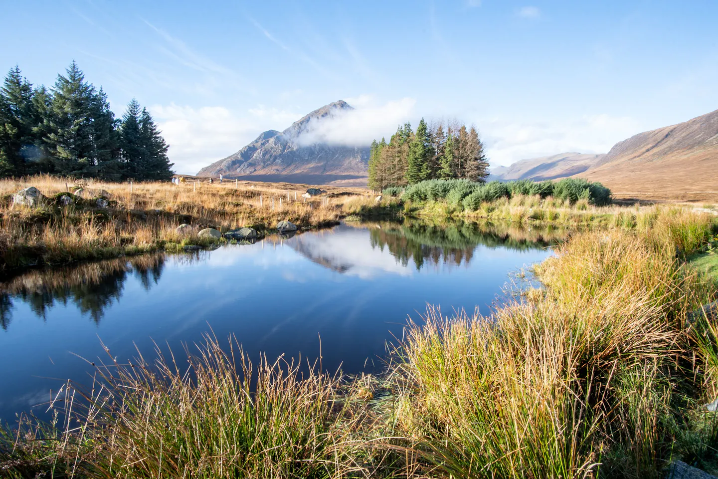 A pyramid-shaped Glencoe mountain wearing a collar of cloud, mirrored in a still dark lochan fringed with golden moor grass and Scots pines