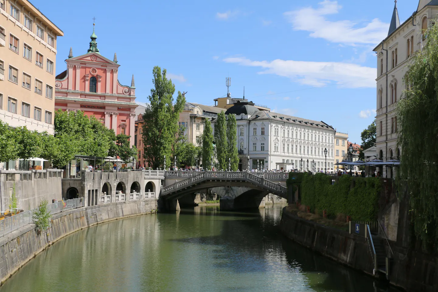 The Triple Bridge over the Ljubljanica in Ljubljana with the pink Franciscan church behind and café umbrellas lining both embankments under a blue sky