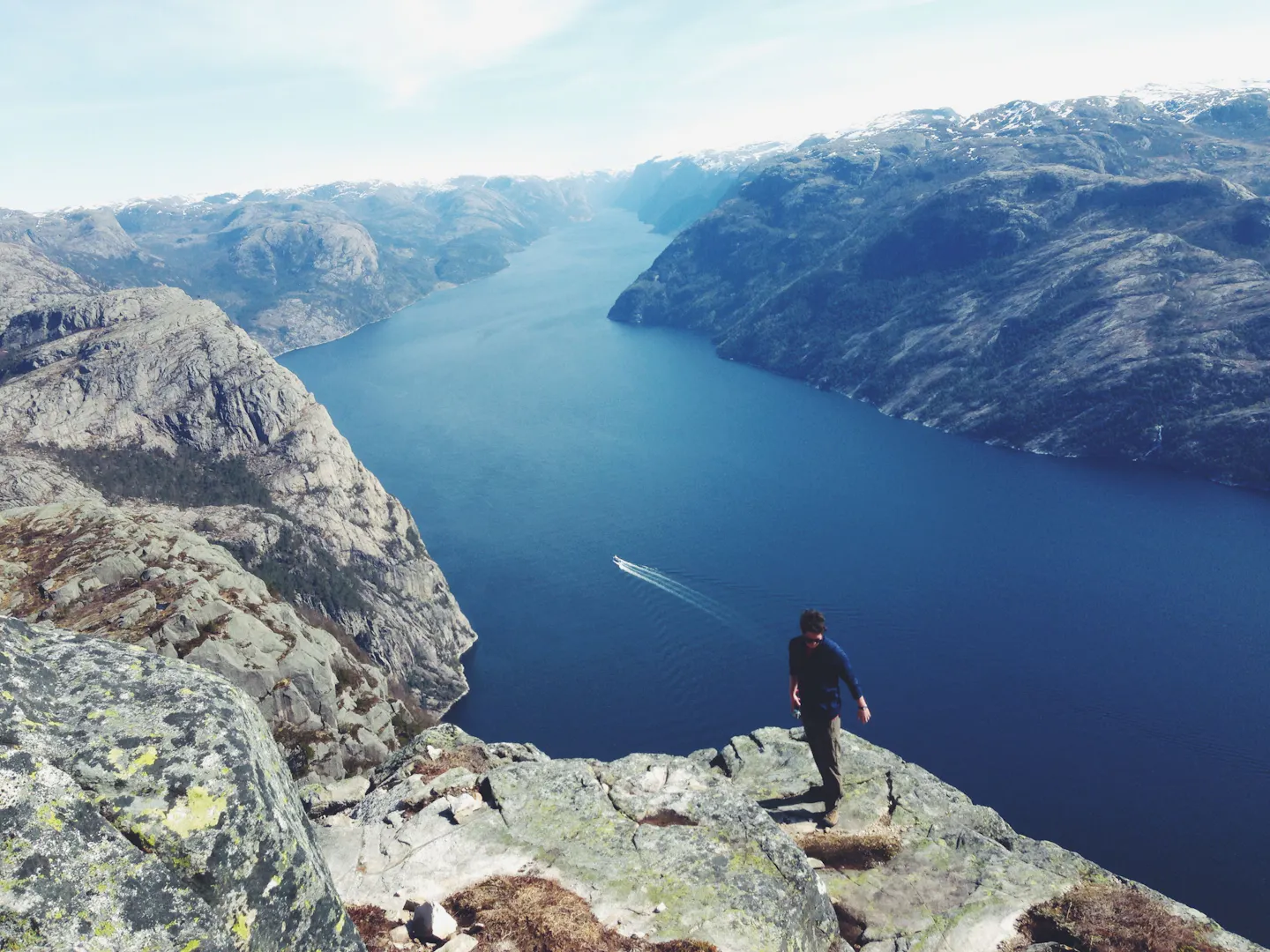 A hiker on a rocky viewpoint high above a deep blue Norwegian fjord, with snow-dusted mountains beyond