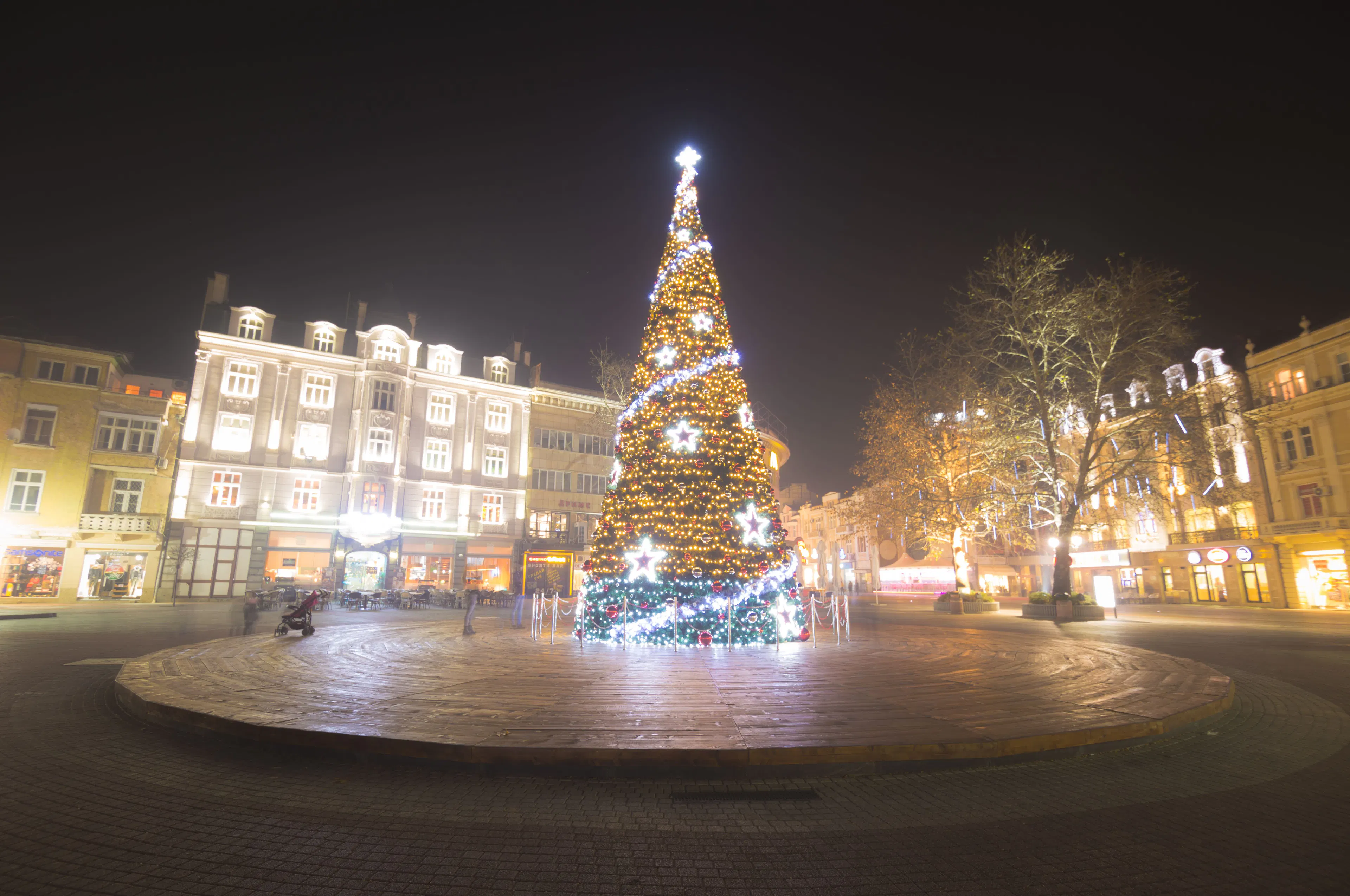 A tall Christmas tree covered in warm lights standing in a European city square at night