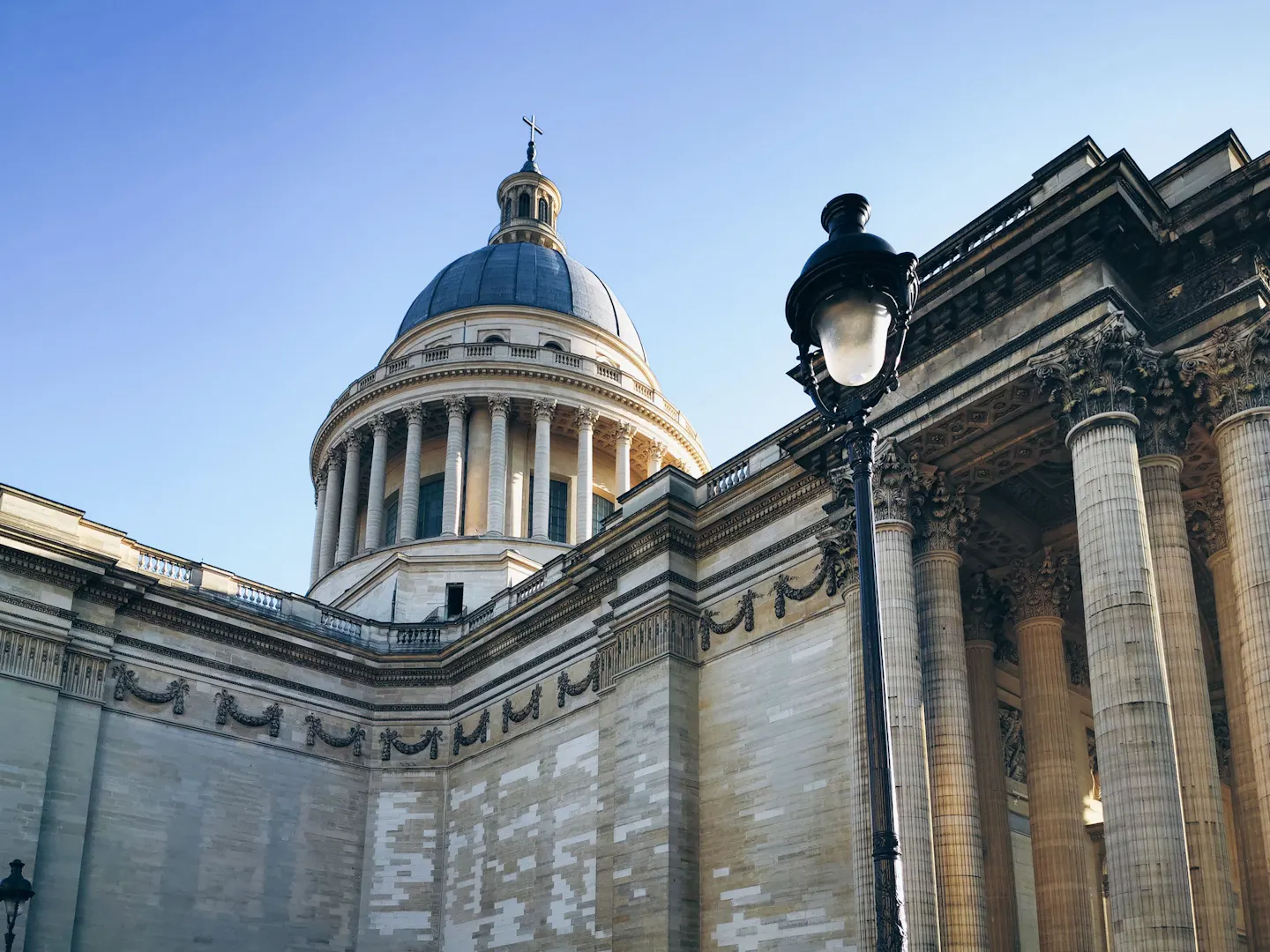 The dome and colonnaded drum of the Pantheon in Paris rising behind the monument's garlanded flank wall, with an ornate cast-iron street lamp in the foreground and a clear blue sky