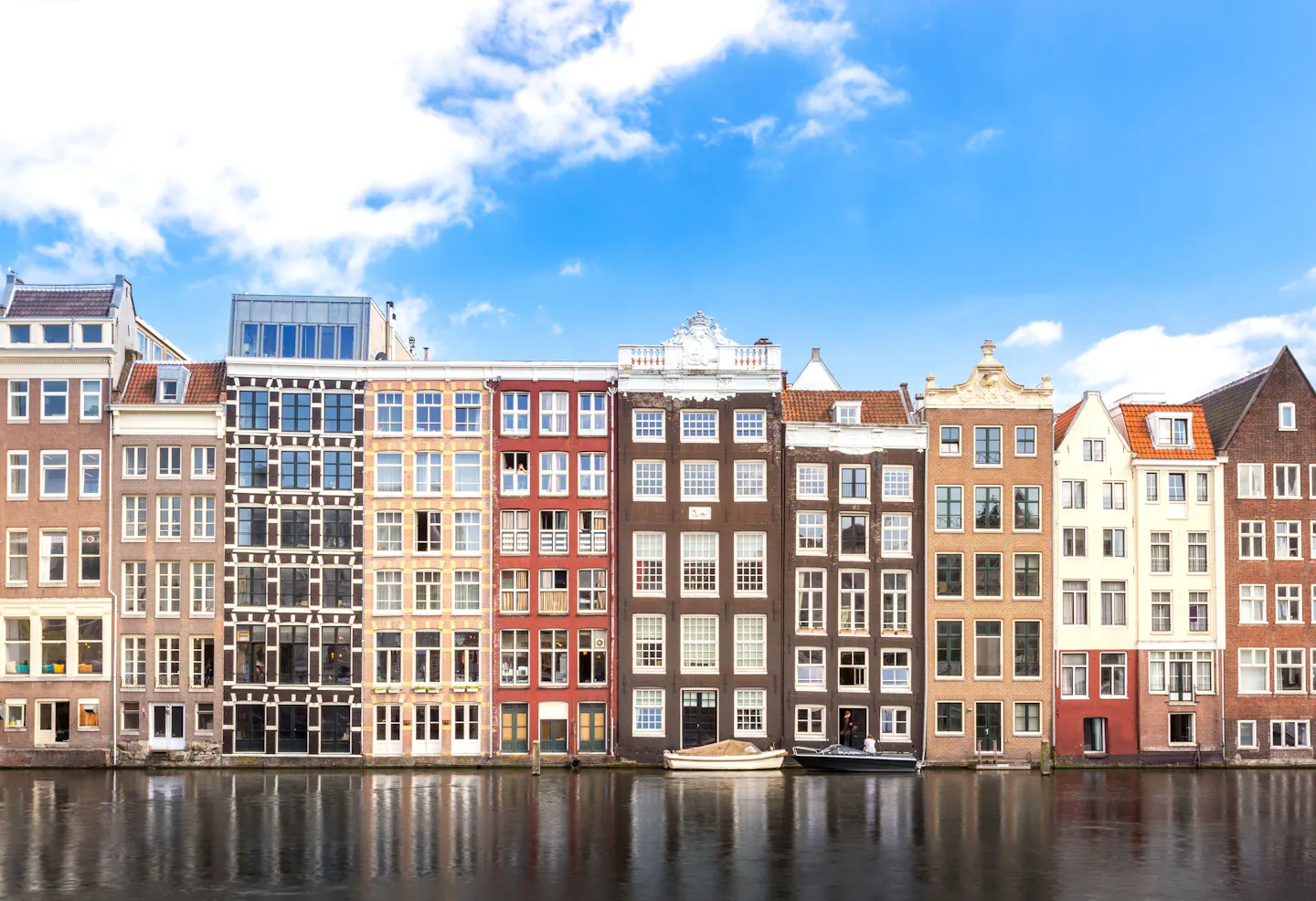 A row of narrow Amsterdam canal houses reflected in still water under a bright blue sky