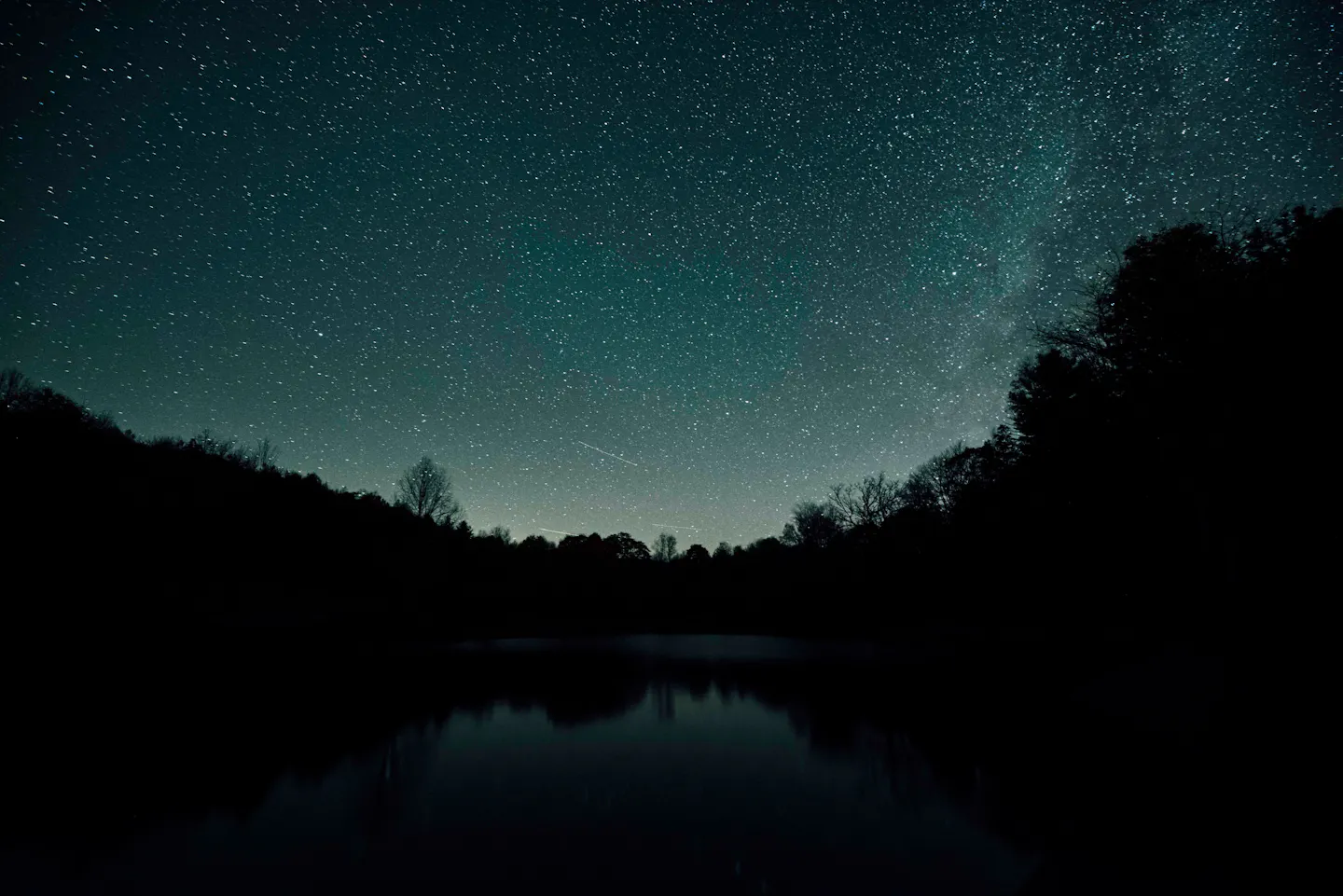 A sky full of stars above the black silhouette of a forest, reflected in the still water of a lake below