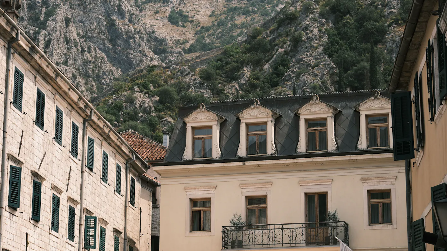 Old-town buildings with green shutters and a balcony beneath steep rocky mountains in Kotor, Montenegro