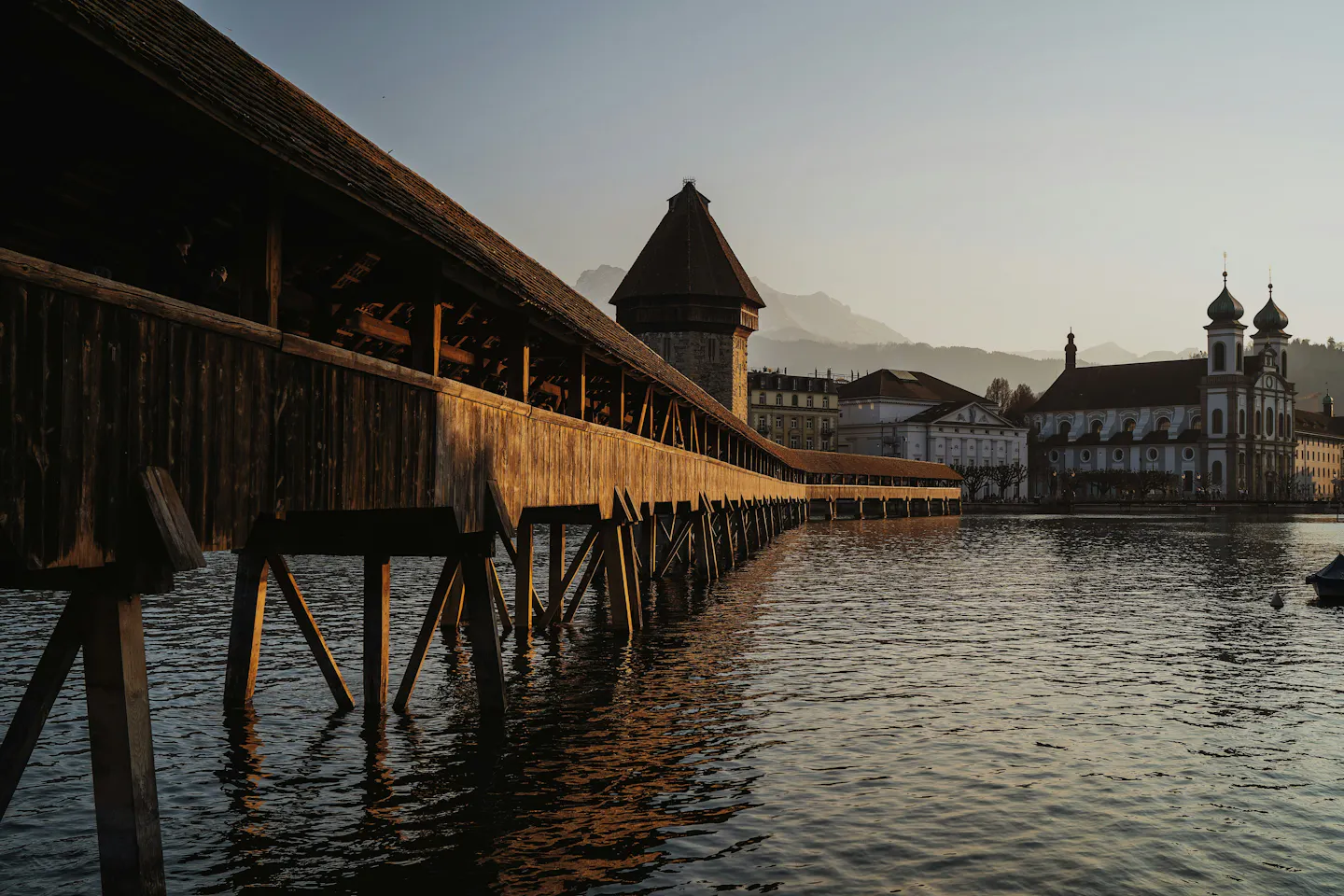 The wooden Chapel Bridge and its octagonal Water Tower on the Reuss at sunset in Lucerne, seen low from the water with golden light along the timber and the old town behind