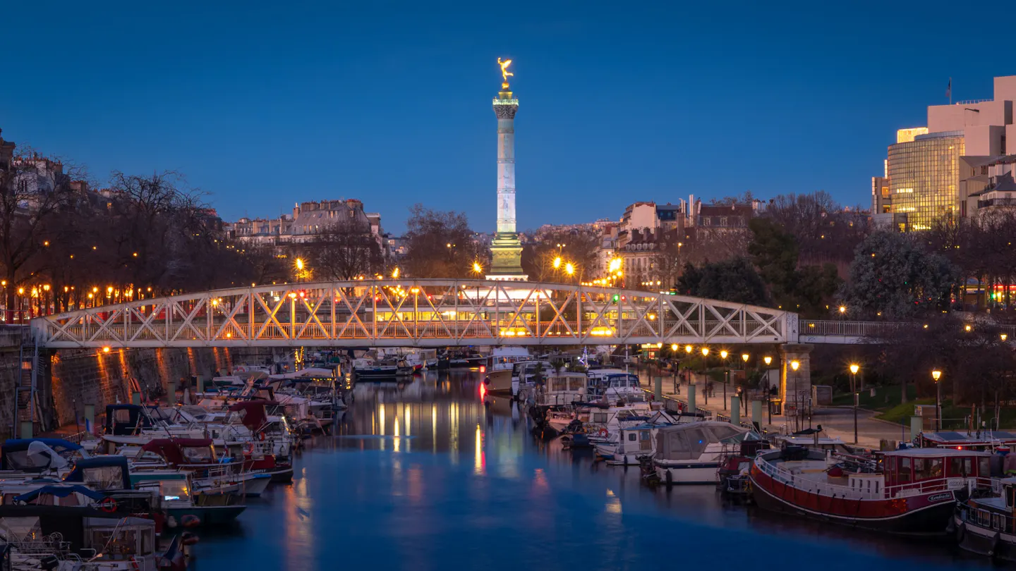 Boats moored along the Port de l'Arsenal at Bastille at dusk, with the lit footbridge and the July Column beyond