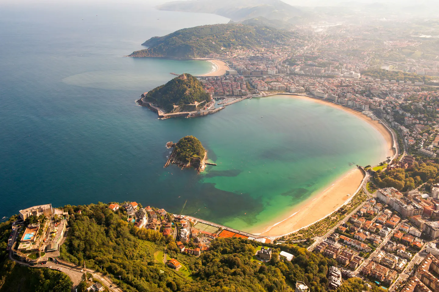 Aerial view of the curved La Concha bay at San Sebastián, with green Santa Clara island in the water and the city wrapped around the sandy crescent