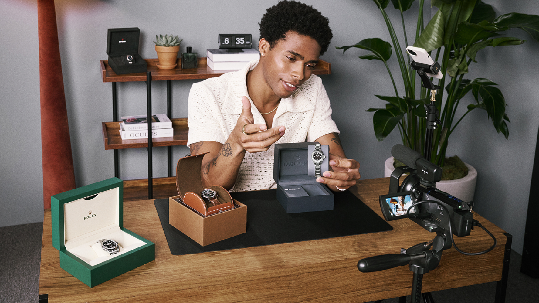 A watch seller records a product video at a desk, holding up a luxury wristwatch to the camera with additional watches and presentation boxes displayed beside him.