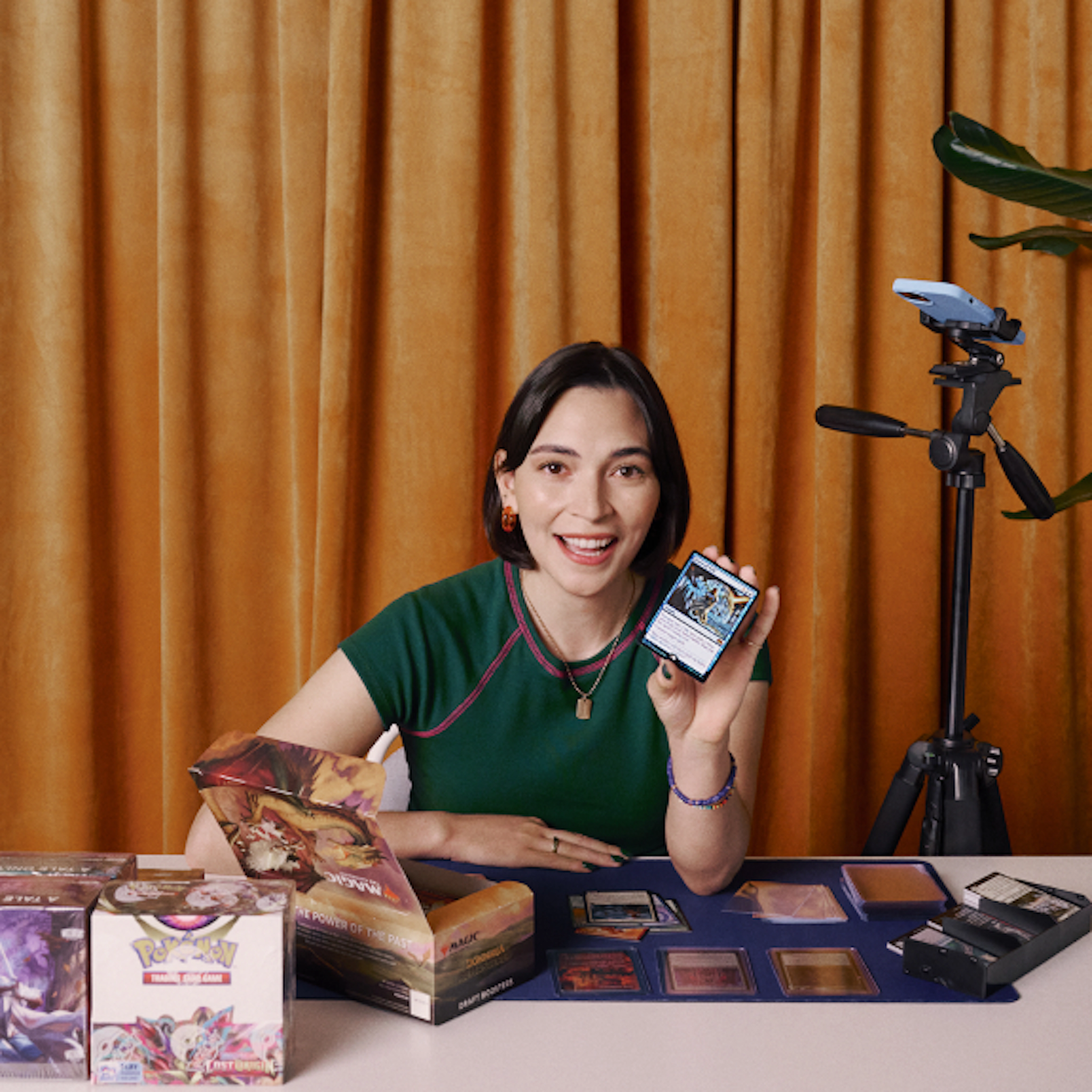 A trading card collector sits at a table with card packs and accessories, smiling while holding up a collectible card beside an overhead camera setup for recording or livestreaming.