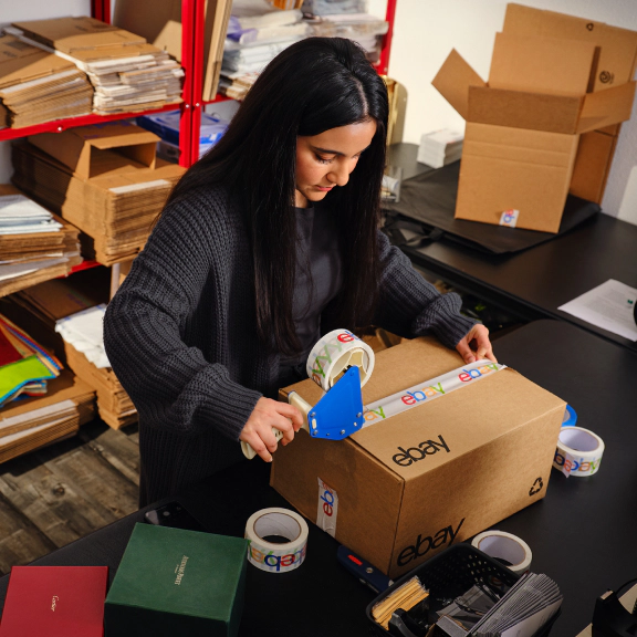 A seller seals an eBay shipping box with branded packing tape at a packing station, surrounded by cardboard boxes, shipping supplies, and packaging materials.