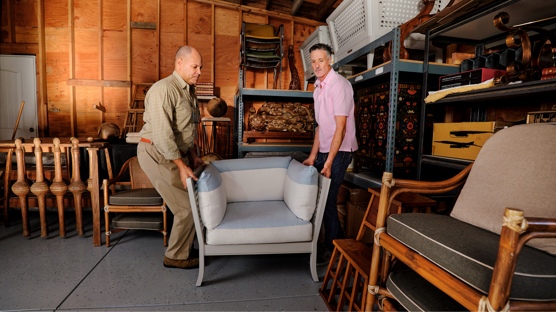 Two furniture sellers carefully carry an upholstered armchair through a warehouse filled with vintage furniture, preparing it for shipping or storage.