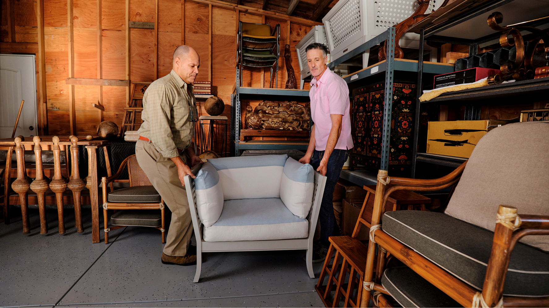 Two furniture sellers carefully carry an upholstered armchair through a warehouse filled with vintage furniture, preparing it for shipping or storage.