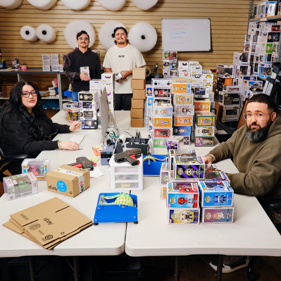 Four collectible Funko Pop sellers work together in a fulfillment space, sorting, packing, and shipping boxed collectibles surrounded by stacks of figurines, shipping supplies, and packing materials.