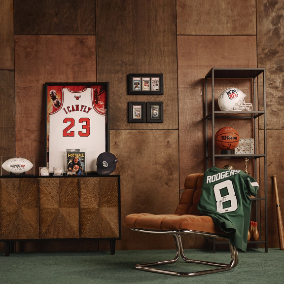 A curated display of sports memorabilia featuring framed jerseys, trading cards, signed footballs, a basketball, and collectibles arranged on furniture and shelving against a wood-paneled wall.
