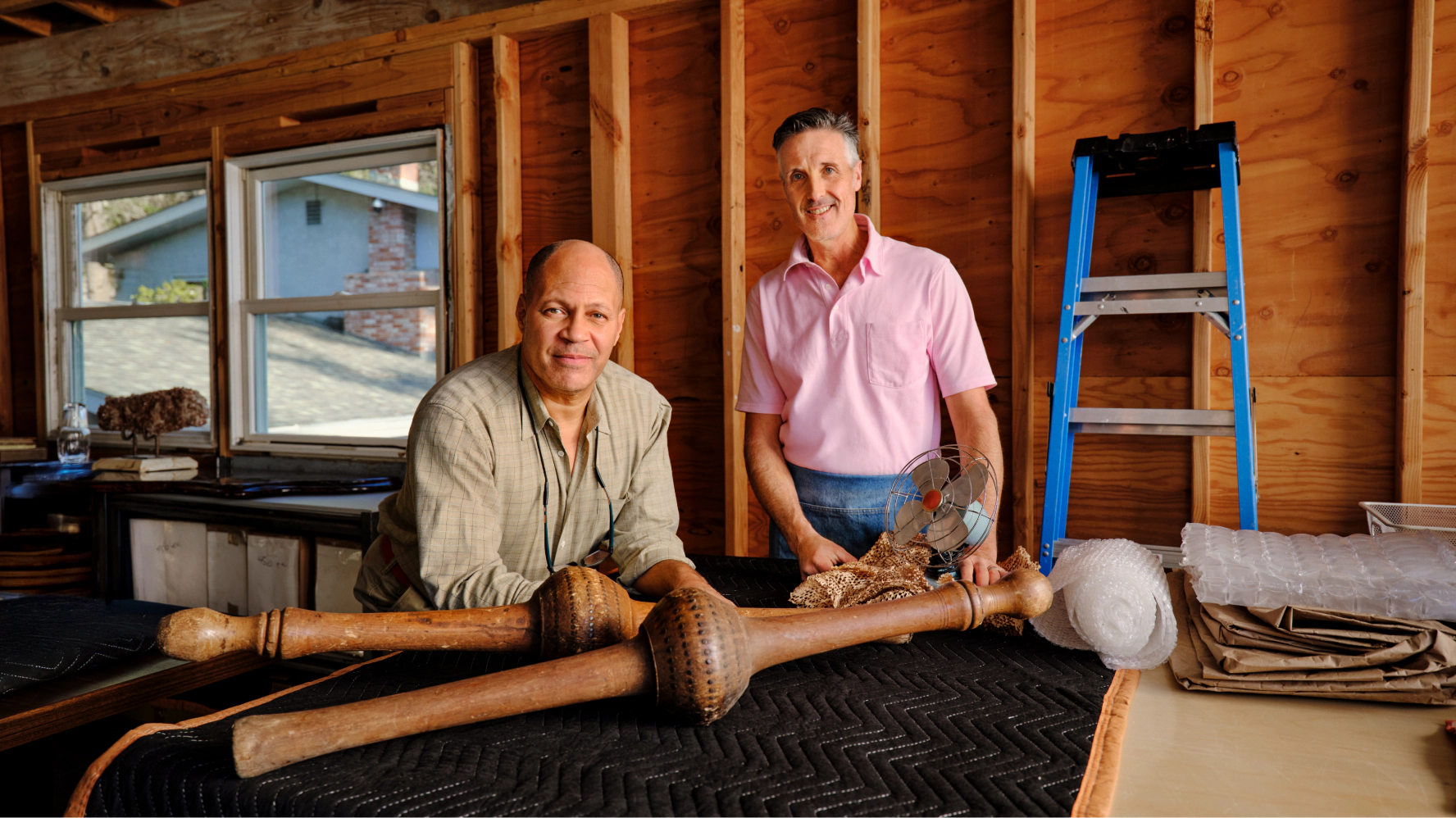 Two antique furniture sellers stand in a workshop preparing vintage furniture for shipment, with carved wooden table legs, packing materials, and moving supplies arranged on a worktable.