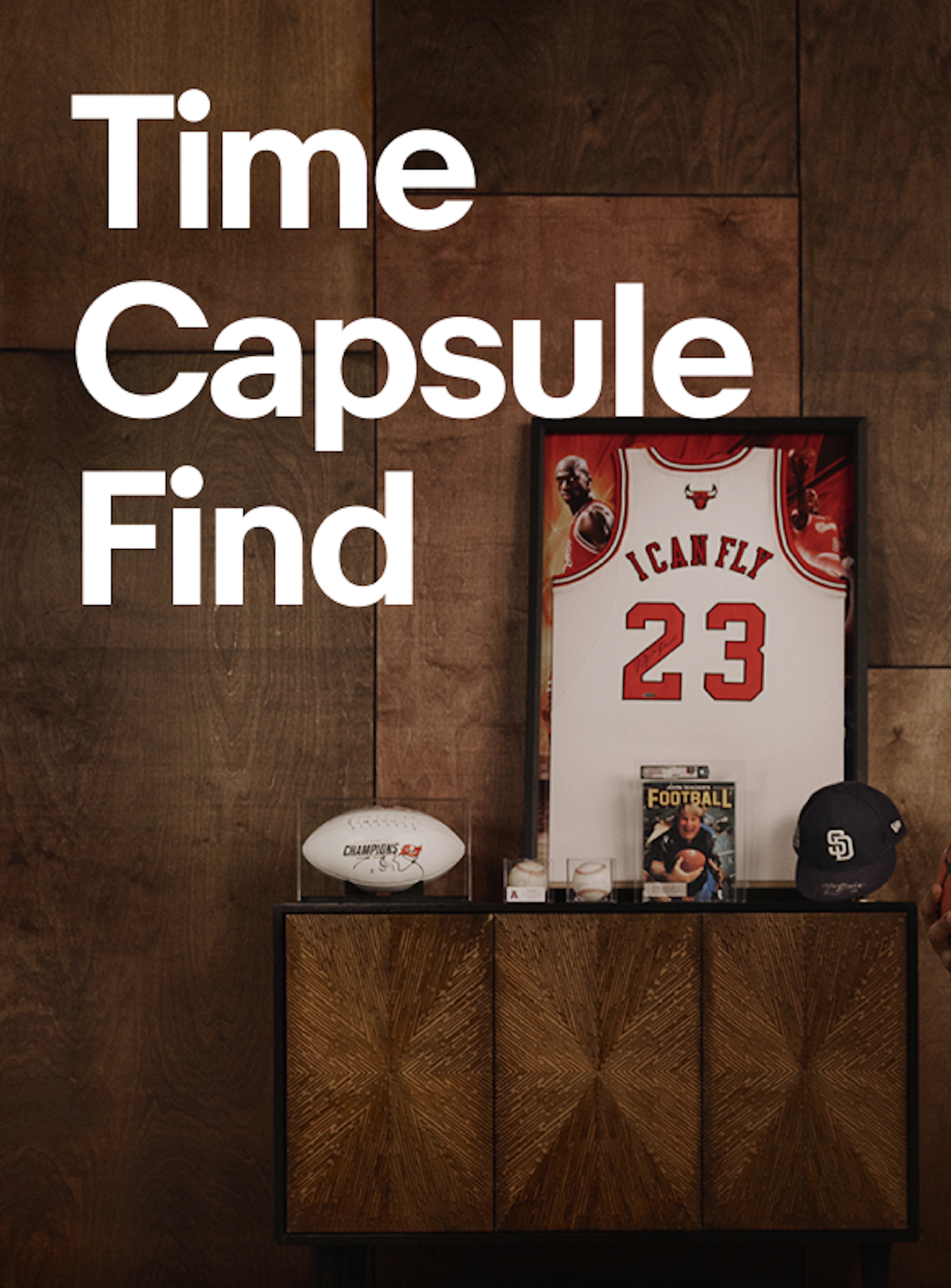 A promotional graphic featuring the headline "Time Capsule Find" above a curated display of sports memorabilia, including a framed basketball jersey, football, baseball cap, magazine, and collectibles arranged on a modern cabinet against a wood-paneled wall.