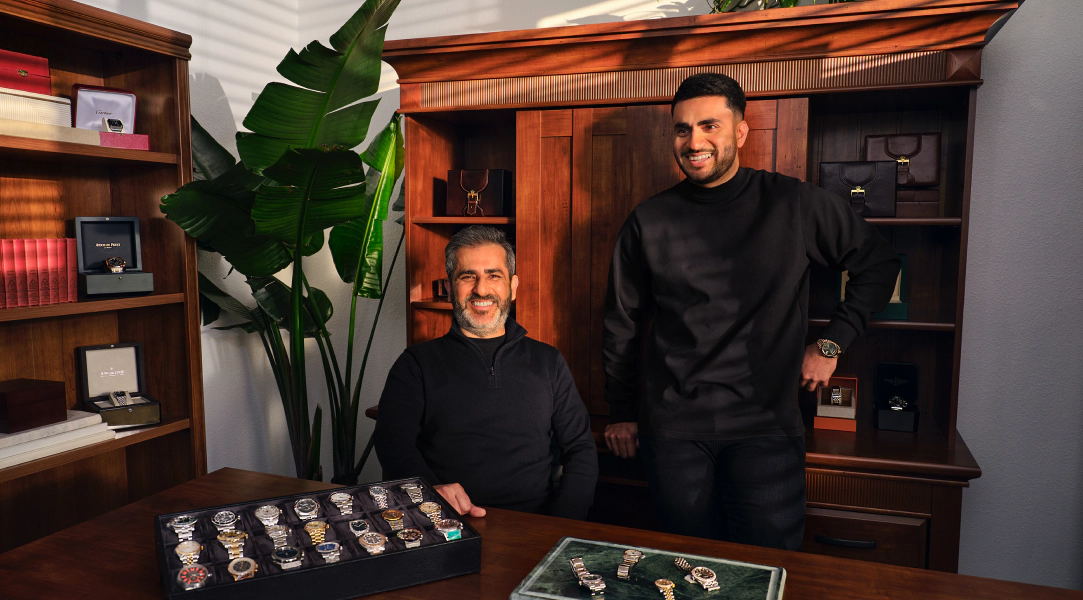 Two watch sellers - father and son - pose in a warmly lit showroom with display trays of luxury watches on a desk and additional timepieces showcased on shelves behind them.