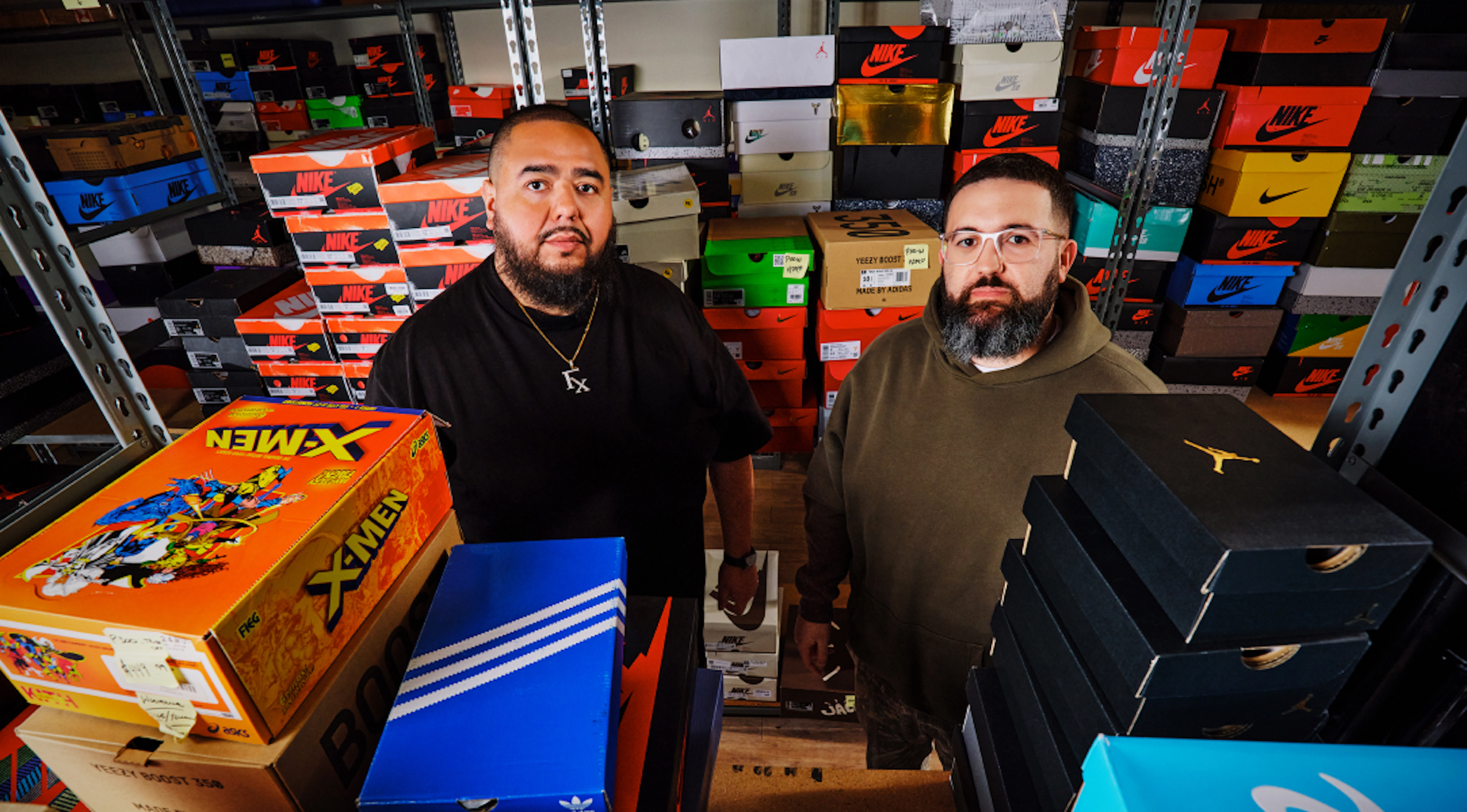 Two sneaker sellers stand in a stockroom surrounded by shelves stacked with shoe boxes from various brands, with additional sneaker boxes in the foreground.