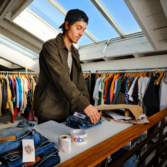 A vintage clothing seller folds and packages a pair of jeans at a packing station, with clothing racks, shipping materials, and eBay-branded packing tape nearby.