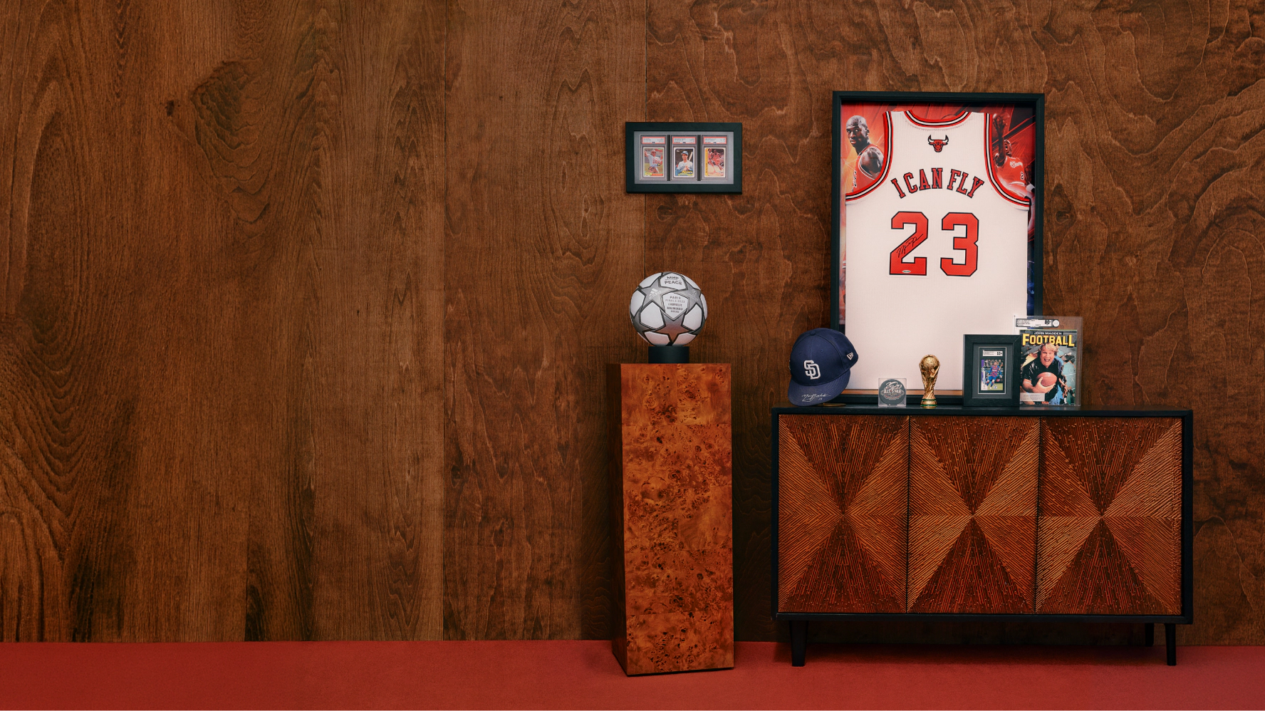 A curated display of sports memorabilia featuring a framed basketball jersey, signed soccer ball, baseball cap, trading cards, magazine, and trophies arranged on a modern cabinet against a wood-paneled wall.