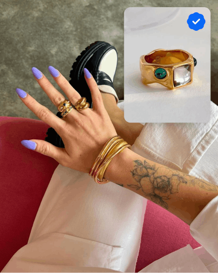 A full bleed image of a woman holding out her hand with purple nails and stacks of gold rings and bracelets. In the upper right corner is a card with a gold, diamond and emerald ring with a blue Authenticity Guarantee badge in the upper right corner.