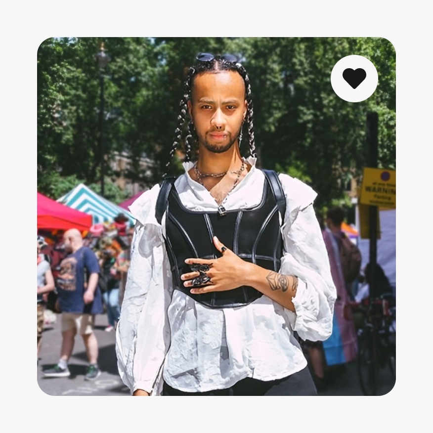 A full bleed lifestyle image of a person with a black corset over a white shirt and braids looking straight faced into the camera. A heart icon is in the upper right corner.