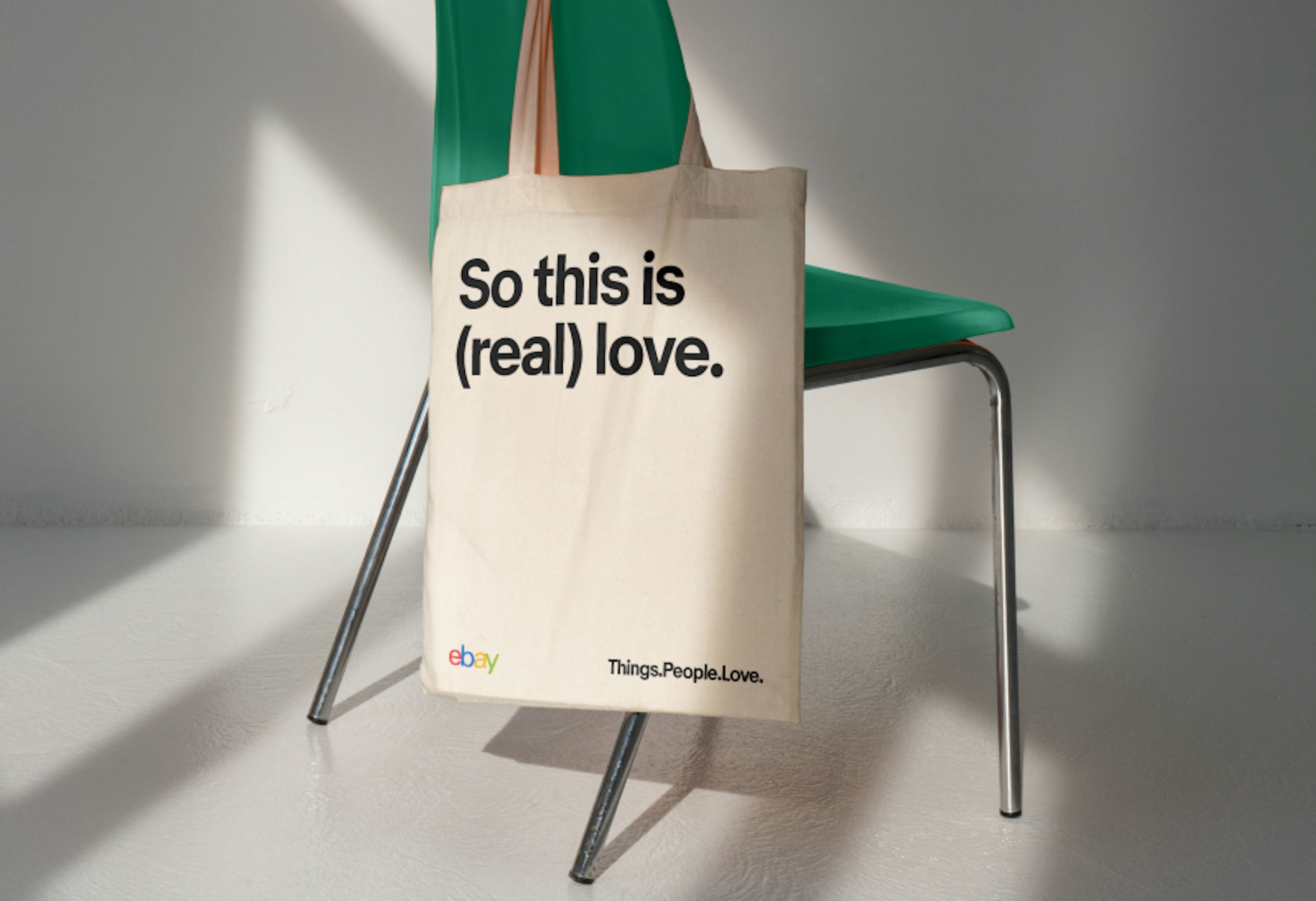 A tote bag that features a bold typographic line, “So this is (real) love.” aligned to the top edge. The logo and tagline lockup are placed along the bottom.
