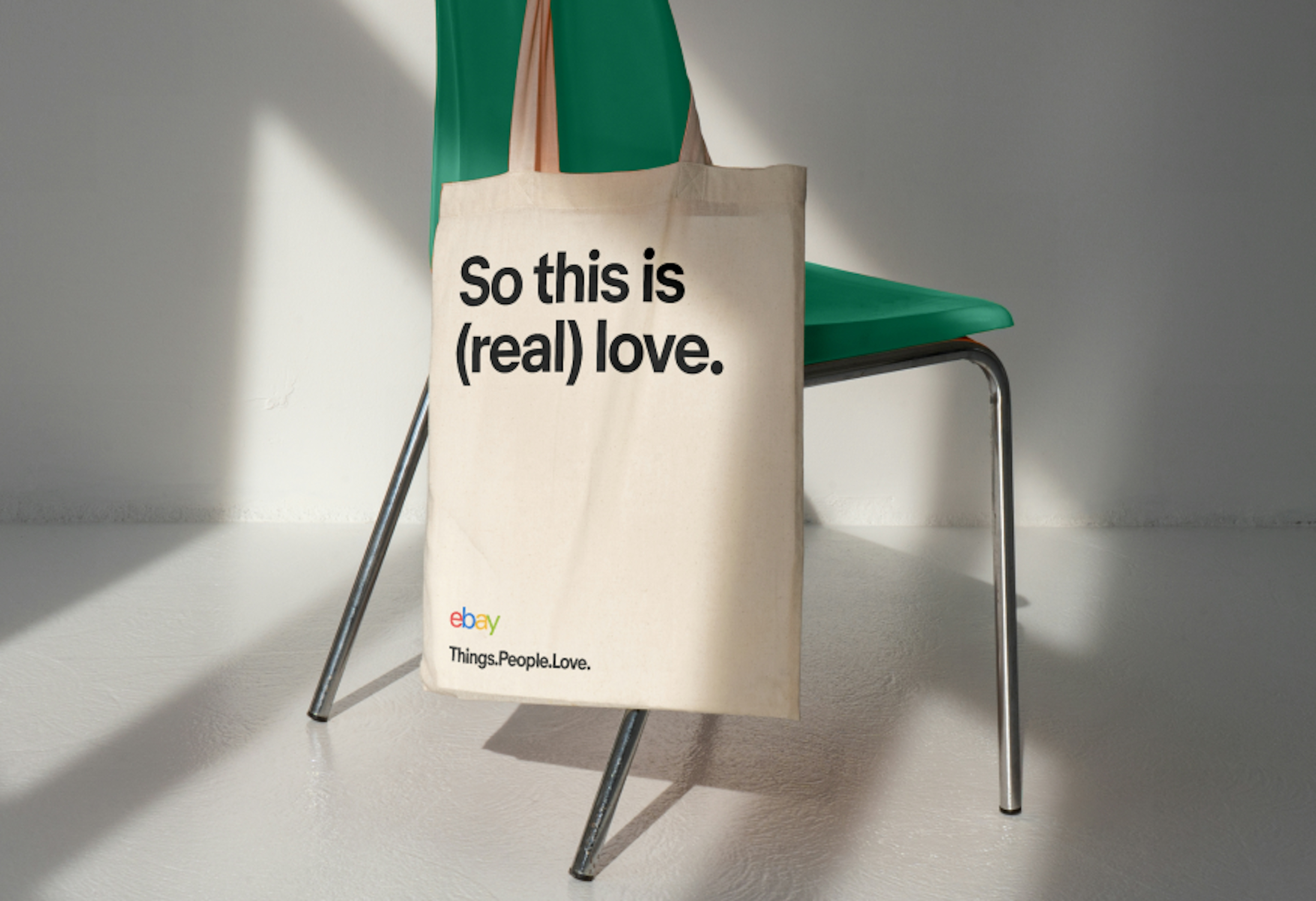 A tote bag that features a bold typographic line, “So this is (real) love.” aligned to the top edge. The logo and tagline lockup is incorrectly stacked in the lower left corner.
