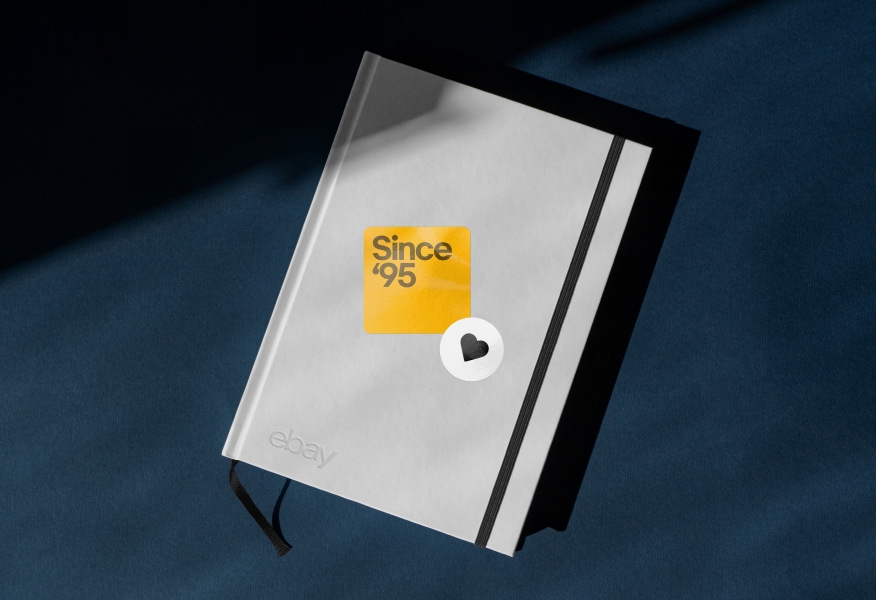 A white notebook with two stickers on the cover: One yellow rounded square with “Since ‘95” set in dark yellow type, and another small white circle with a centered black heart icon.