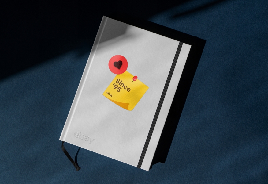 A white notebook with two stickers on the cover: One yellow square with a graphic fold to imitate a sheet of paper with a graphic red push pin, and another small red circle with a centered red icon with a drop shadow applied as a 3D effect.
