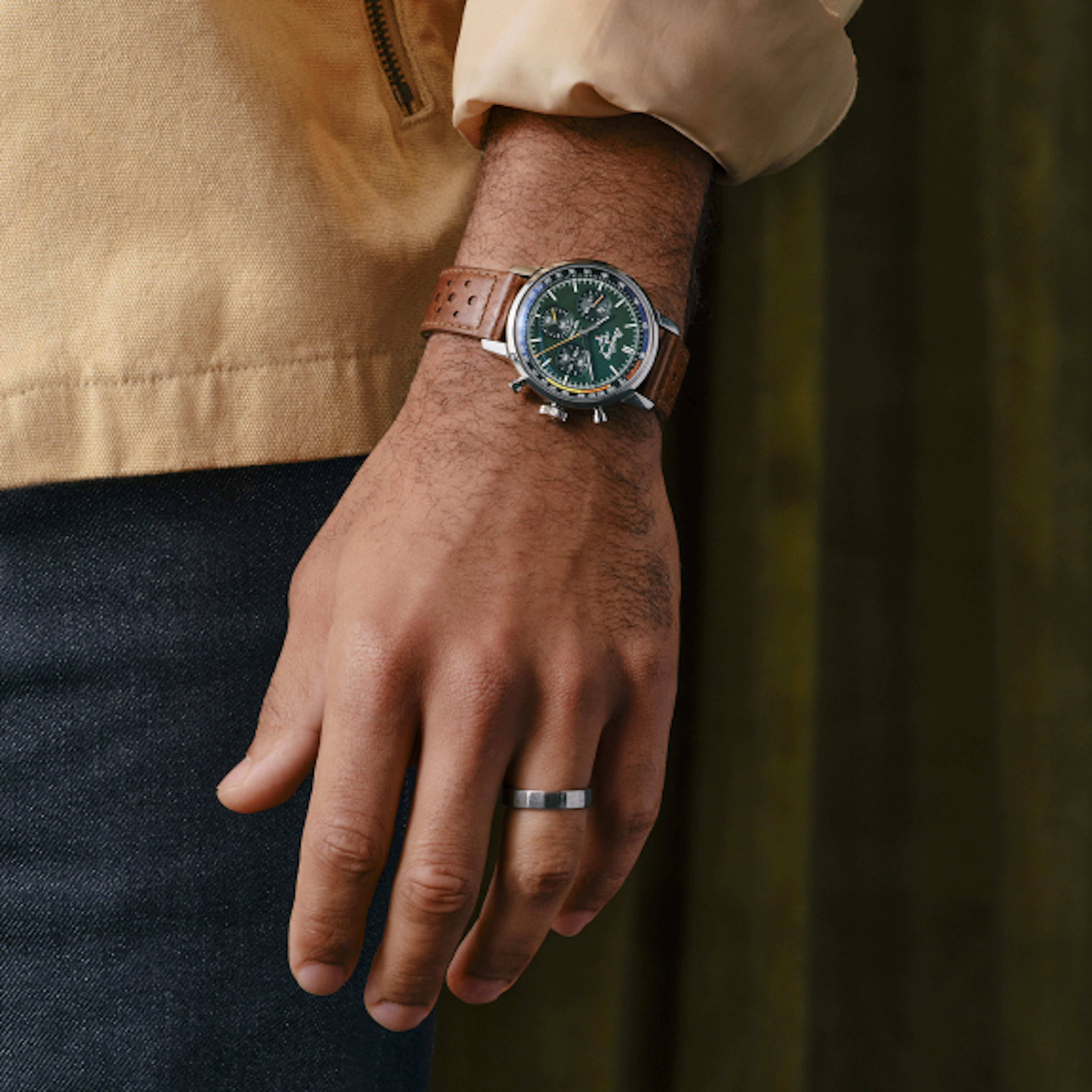A close-up of a person's hand wearing a luxury wristwatch with a brown leather strap and green dial, alongside a silver ring on their finger.