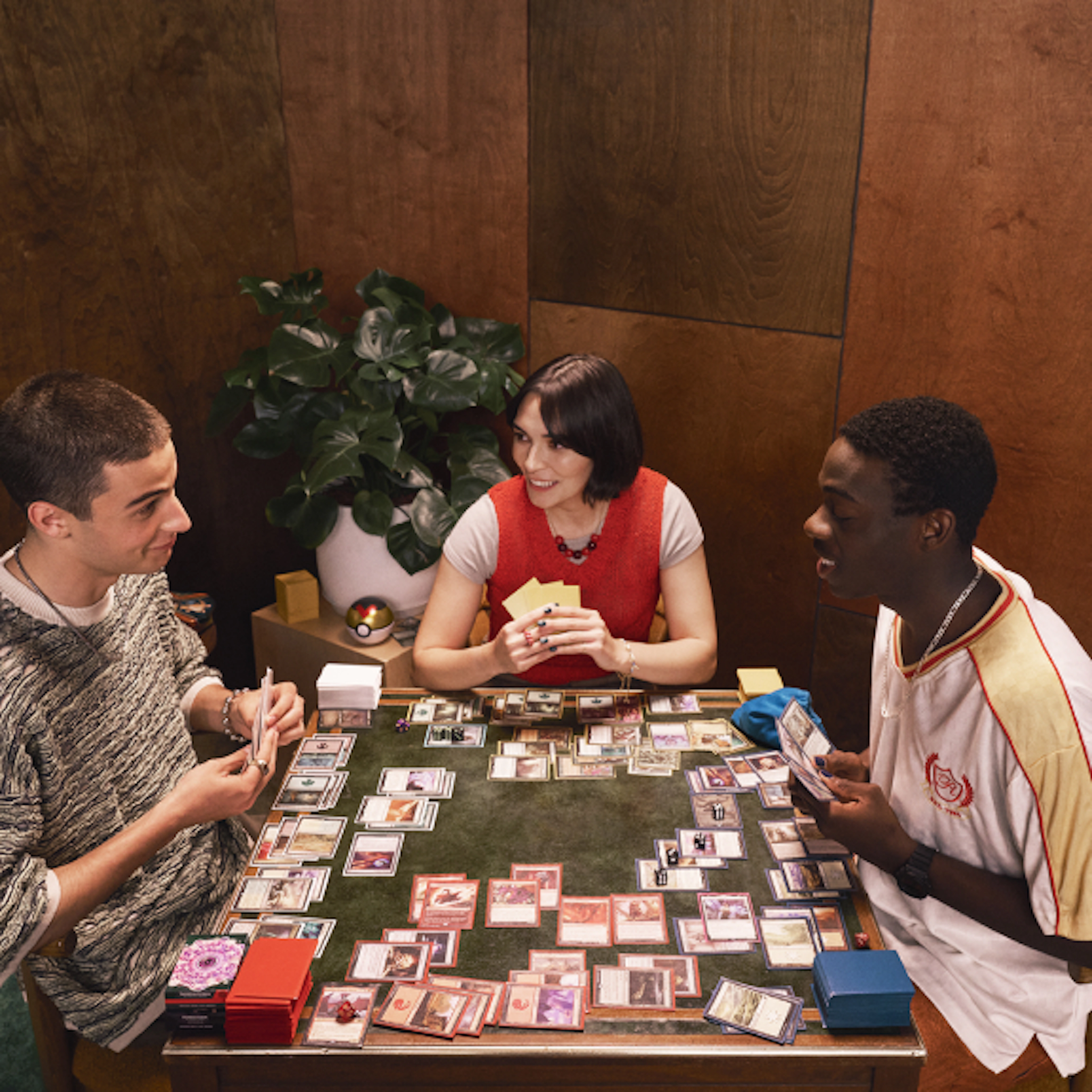 Three people sit around a table playing a trading card game, holding cards and smiling as a game in progress is spread across the tabletop.